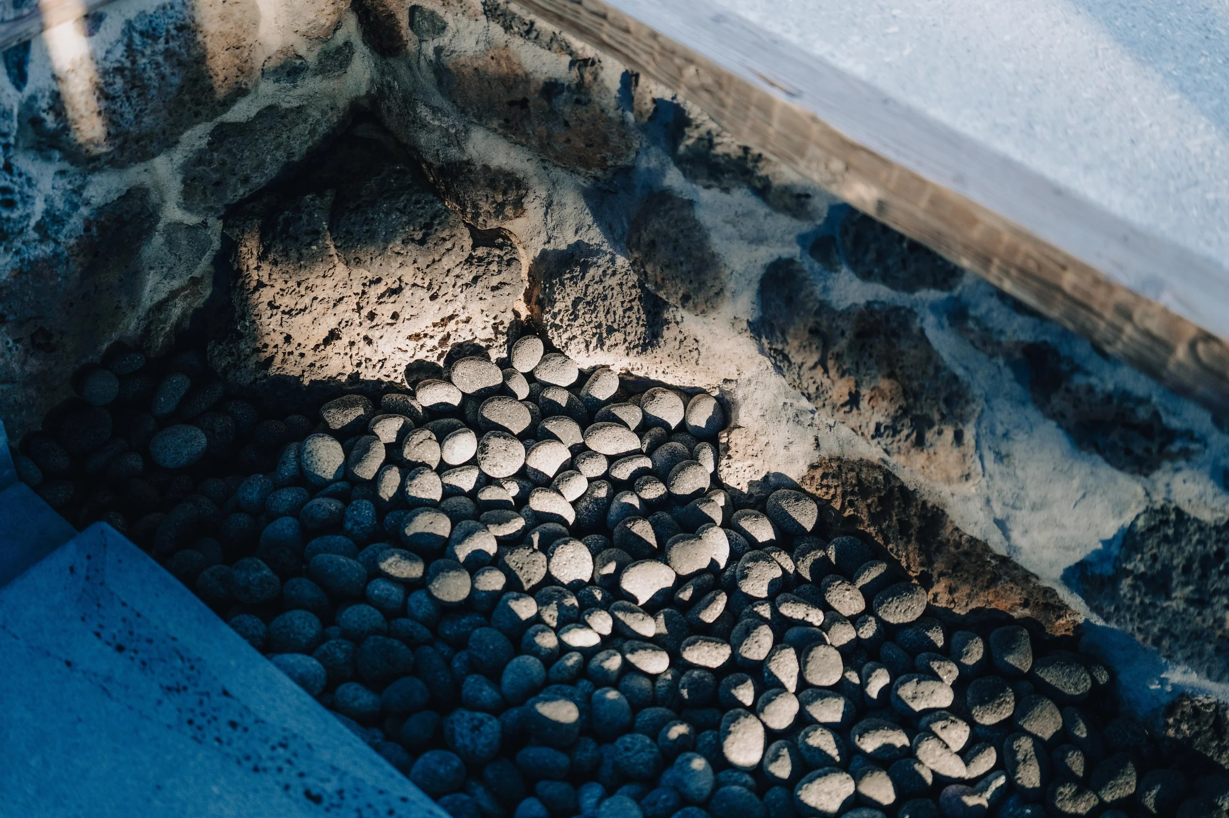 NUUT Aewol 눗애월 Close-up view of small, smooth, black stones within a stone-lined fireplace or hearth area, with some light illuminating the stones and textured rocks.