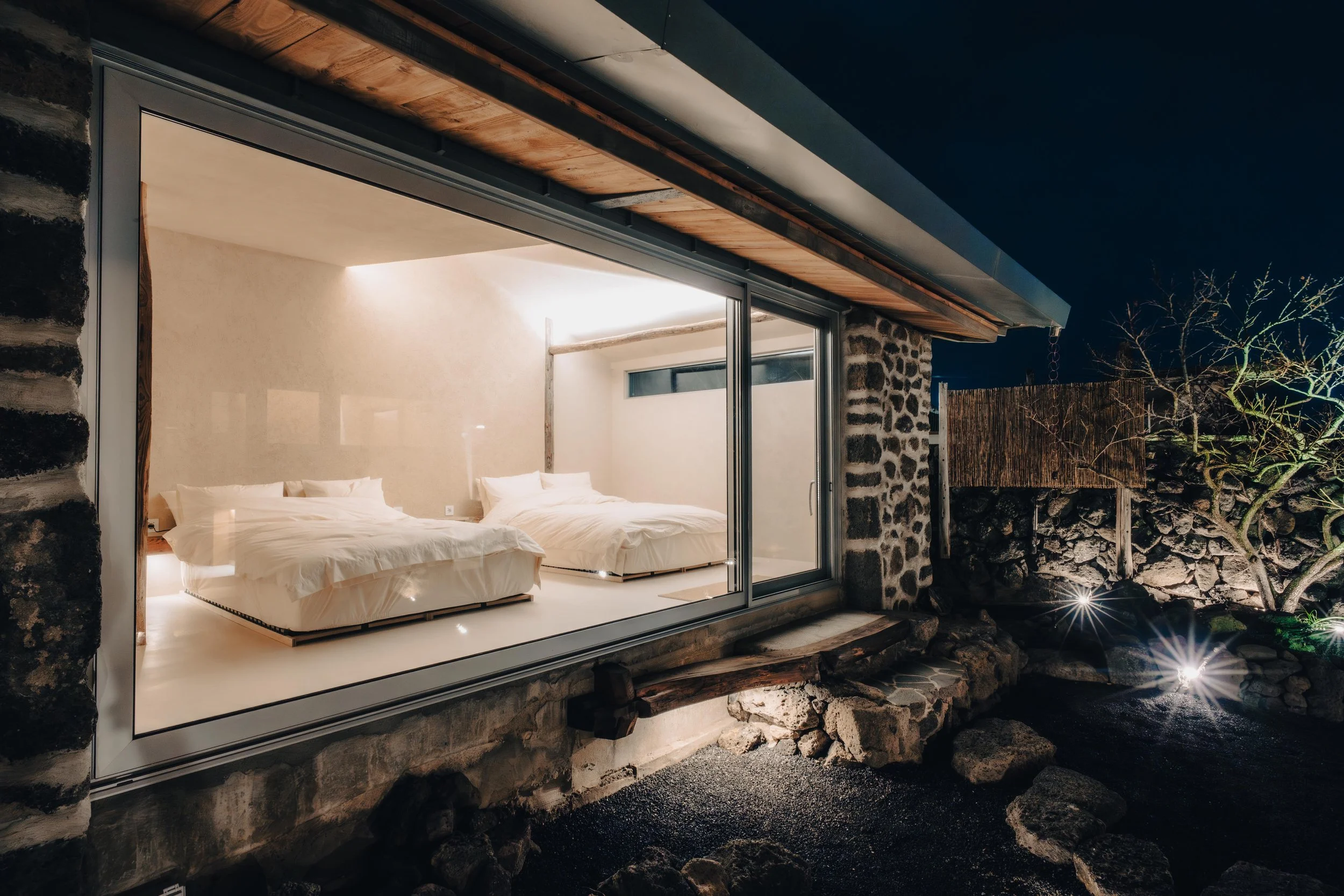 NUUT Aewol 눗애월 Night view of a modern house with large glass windows revealing a bedroom with two beds inside. The house features stone and wood accents, and outdoor lighting illuminates rocks and a tree outside.