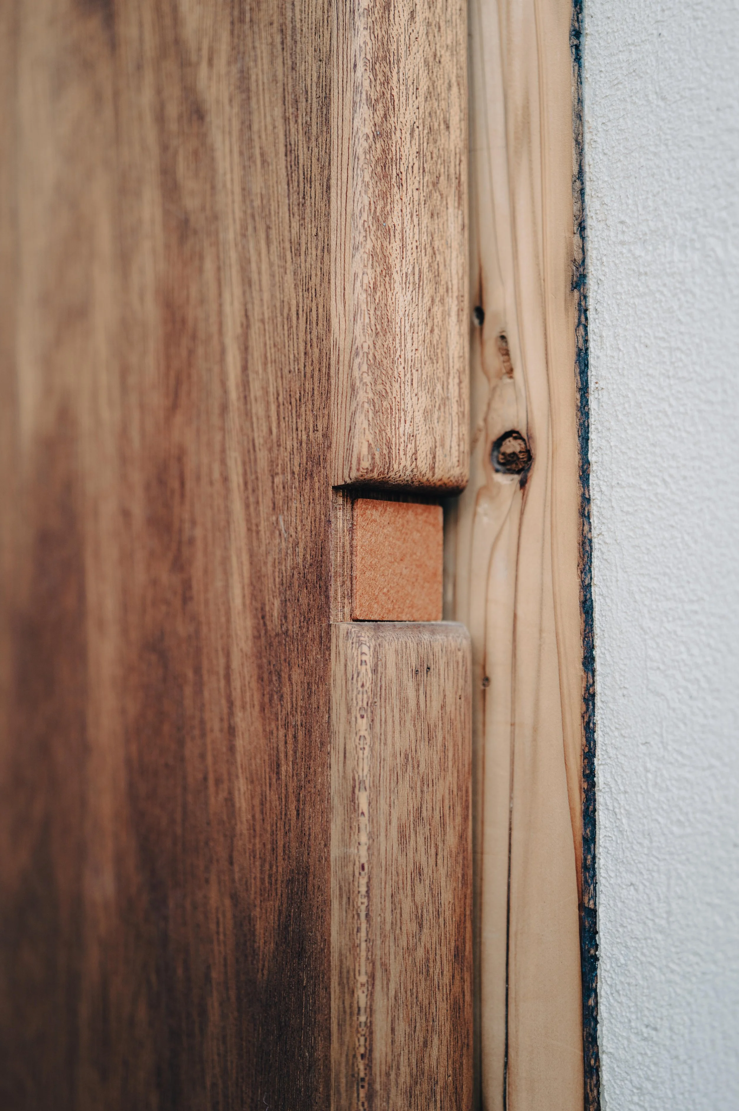 NUUT Aewol 눗애월 Close-up of a wooden door with a small brick piece inserted in a gap near the edge, adjacent to a textured white wall.
