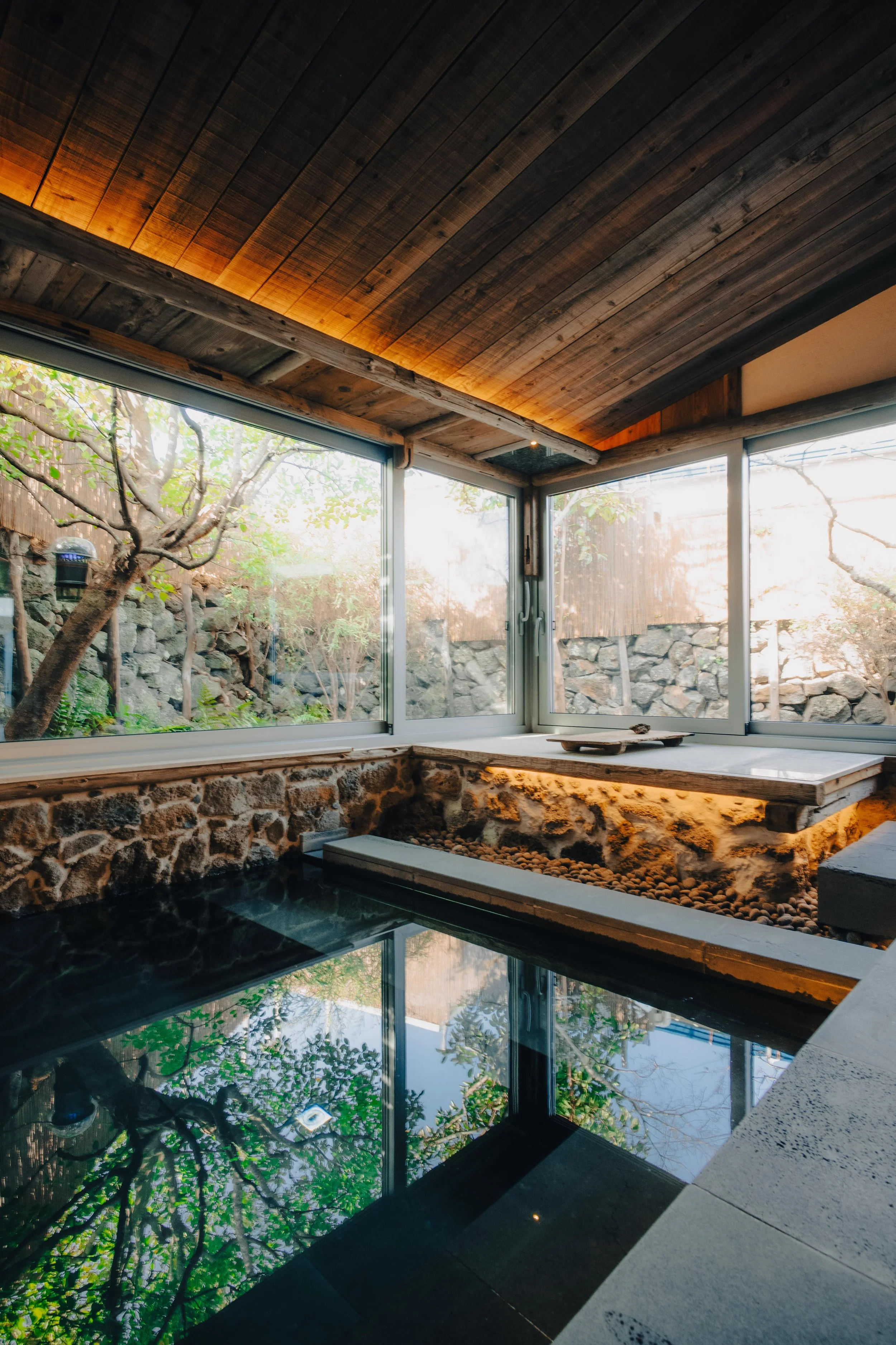 NUUT Aewol 눗애월 Indoor hot spring bath with large windows showing trees and rocks outside, wooden ceiling, and stone and tile walls.