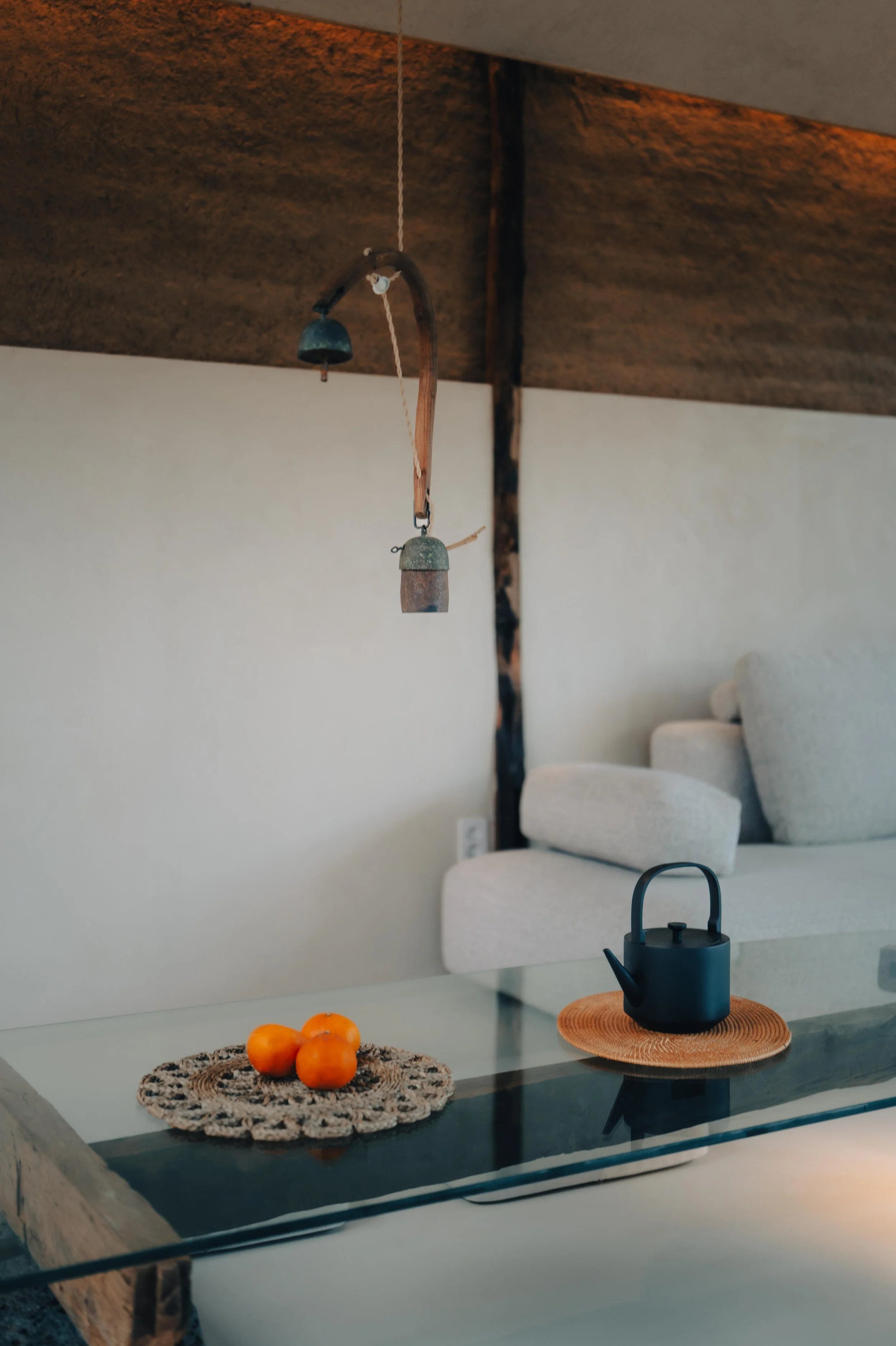 NUUT Aewol 눗애월 A black teapot on a round woven placemat on a glass table, with three oranges on a crocheted doily, and a rustic wind chime hanging from the ceiling in a cozy living room.