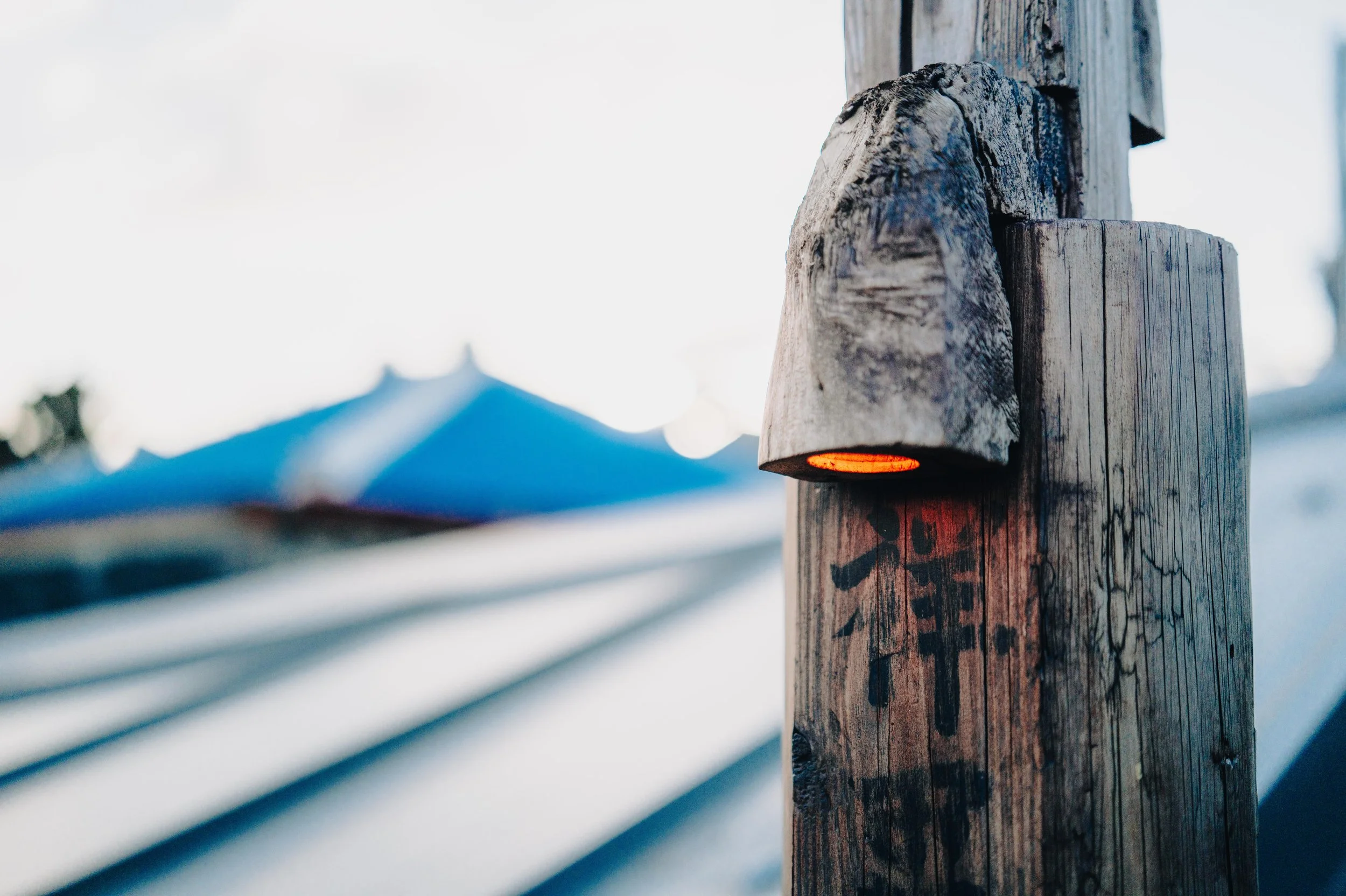 NUUT Aewol 눗애월 Close-up of wooden post with Japanese writing and small red light, blurred background of blue umbrellas and sky.