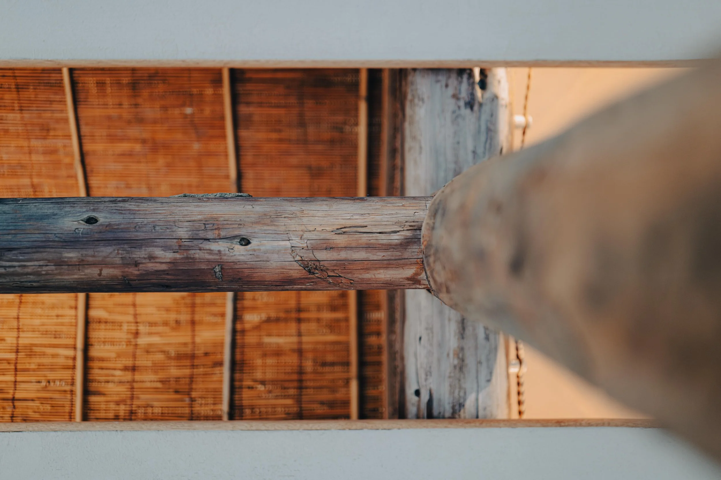 NUUT Aewol 눗애월 Close-up of weathered wooden posts supporting a structure with a ceiling made of bamboo or wooden slats, sunlight illuminating the scene.