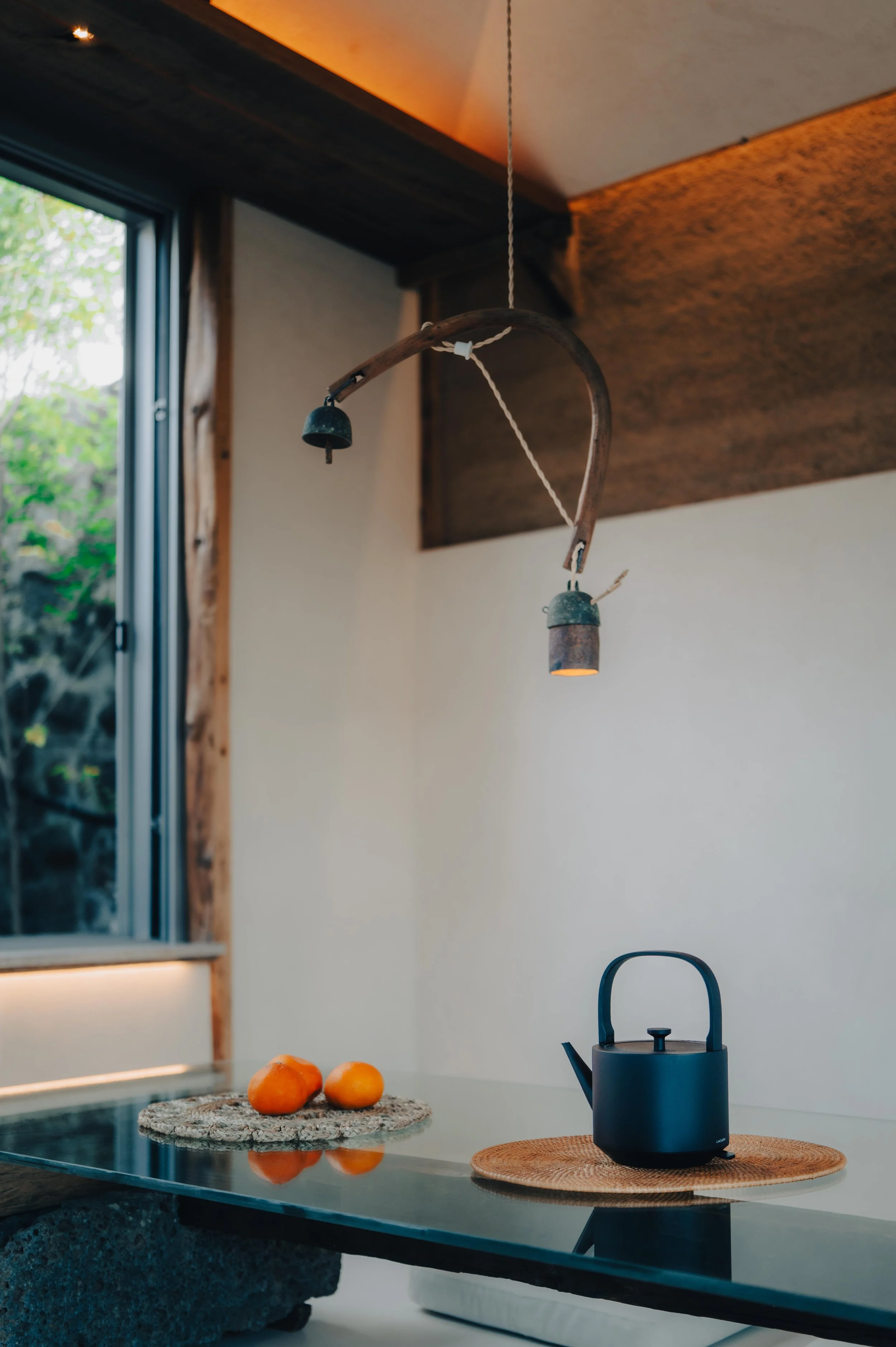 NUUT Aewol 눗애월 A black electric kettle on a woven placemat, three oranges on a small rug, and a window with greenery outside in a cozy, rustic kitchen setting.