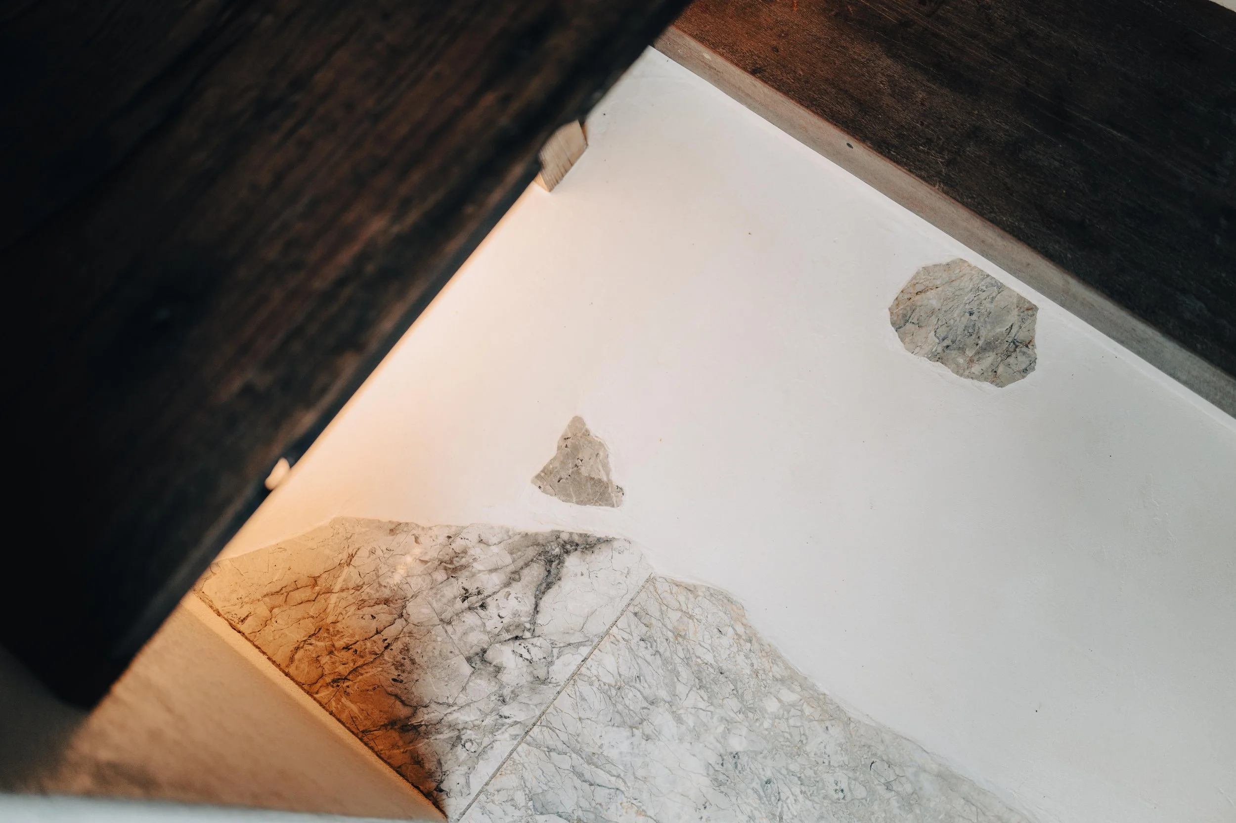NUUT Aewol 눗애월 View of a corner of a room showing part of a dark wooden piece of furniture, a textured white wall with two patches of damaged plaster, and a marble floor.