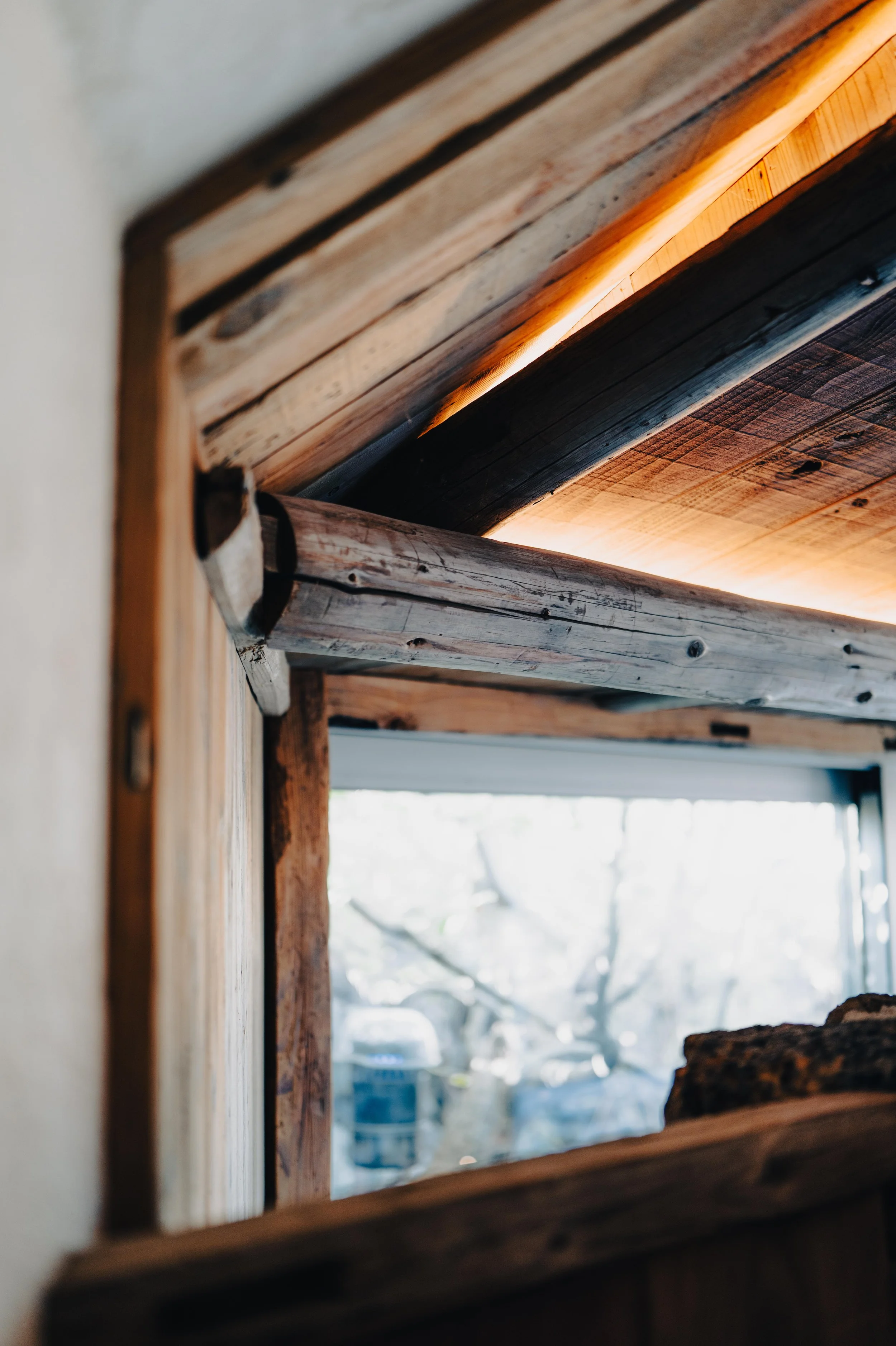 Close-up of a wooden window frame and interior ceiling with exposed wooden beams, looking outward through the window to trees and outdoor objects.