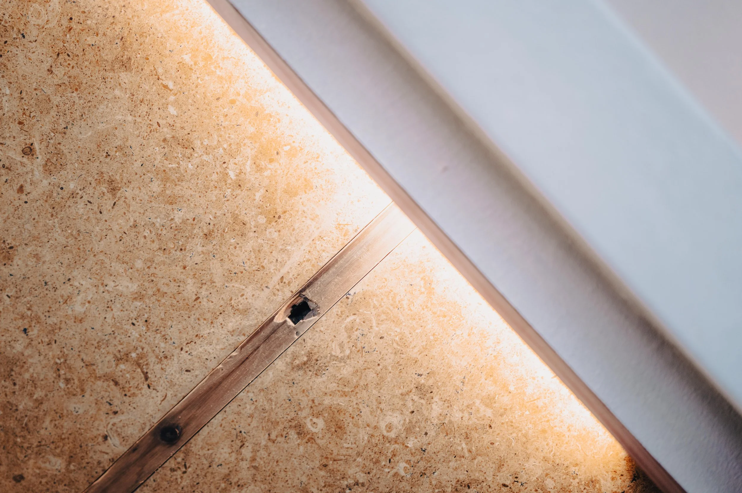 NUUT Aewol 눗애월 Close-up of a gap under a light-colored cabinet with a strip of LED lighting on the underside, revealing a damaged baseboard and small hole in the wood trim along the floor.
