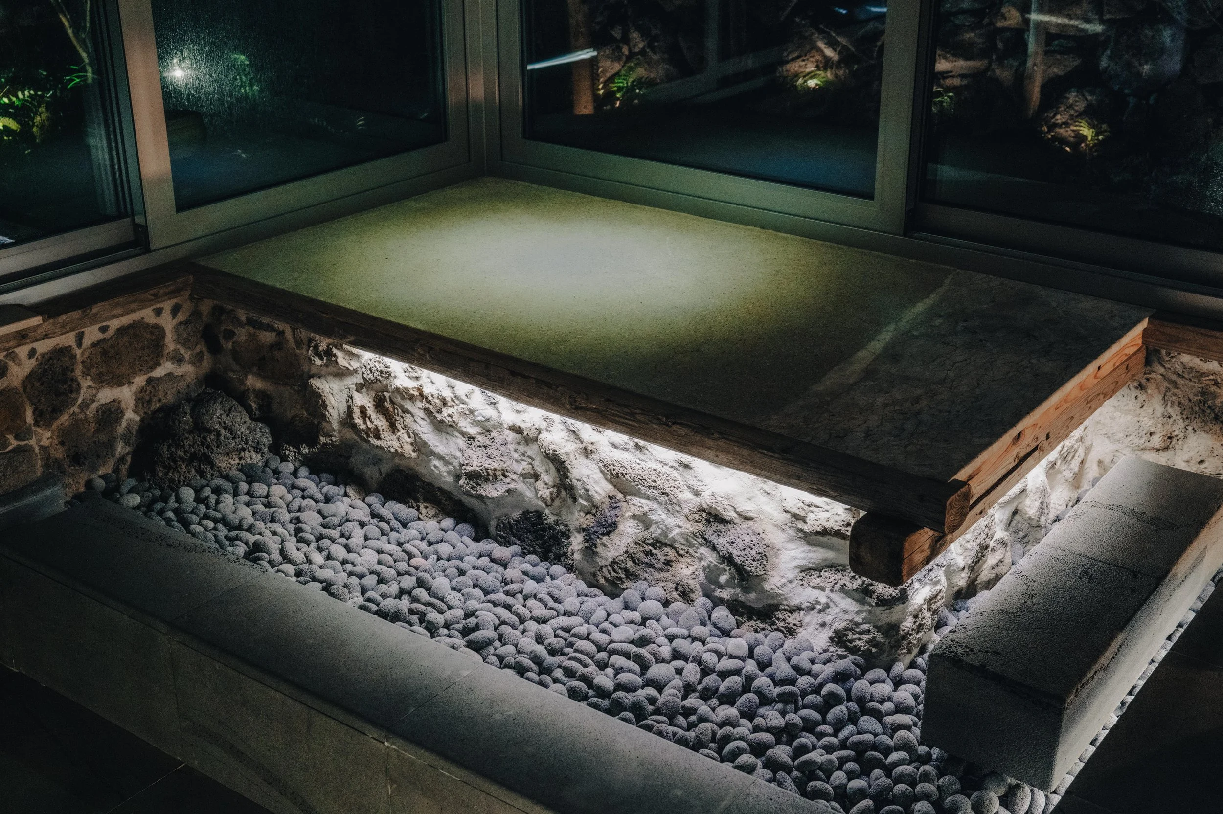 NUUT Aewol 눗애월 Indoor space with a glass enclosure, a concrete ledge with a stone and pebble base, and a patch of green grass on top of a wooden frame.