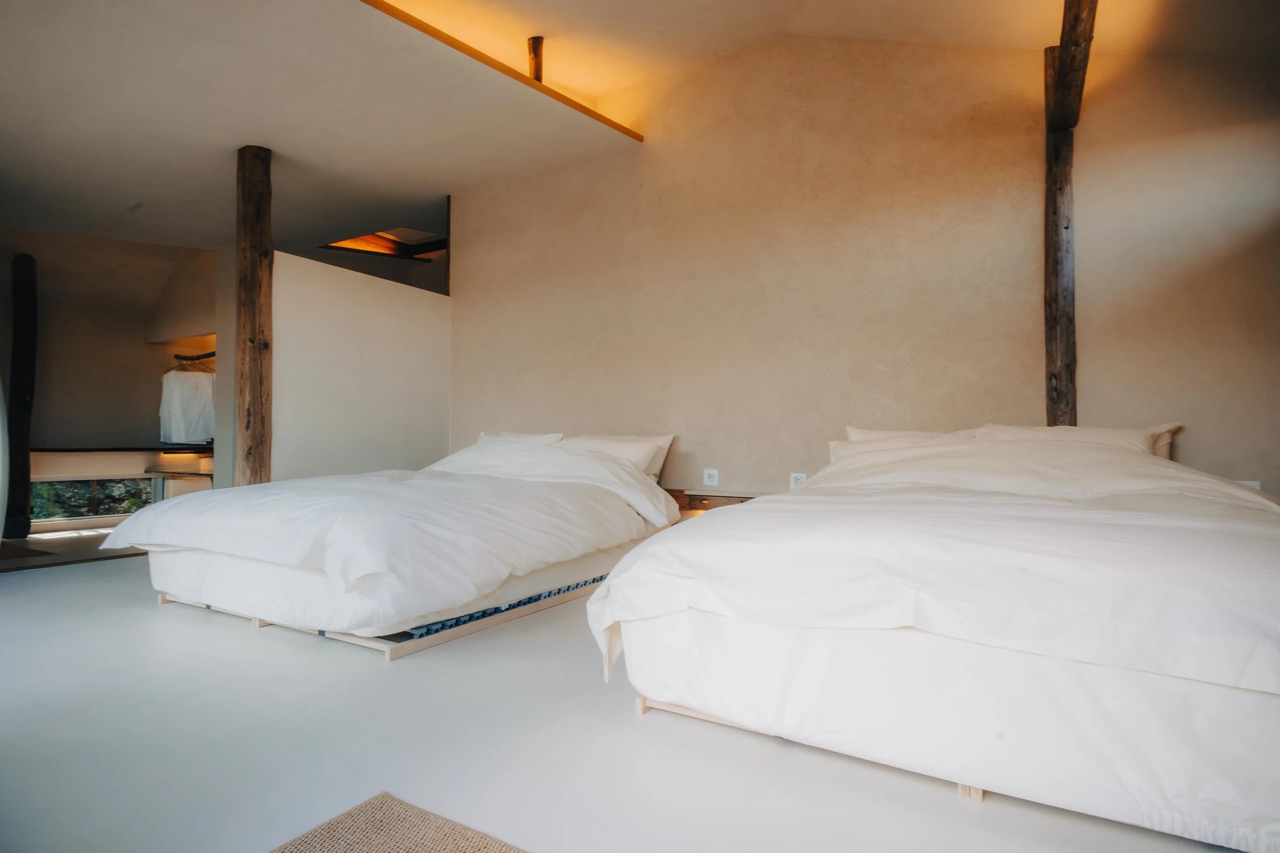 NUUT Aewol 눗애월 Two neatly made beds with white bedding in a modern, minimalist bedroom with natural wood beams and beige walls, soft lighting, and a small window on the left side.