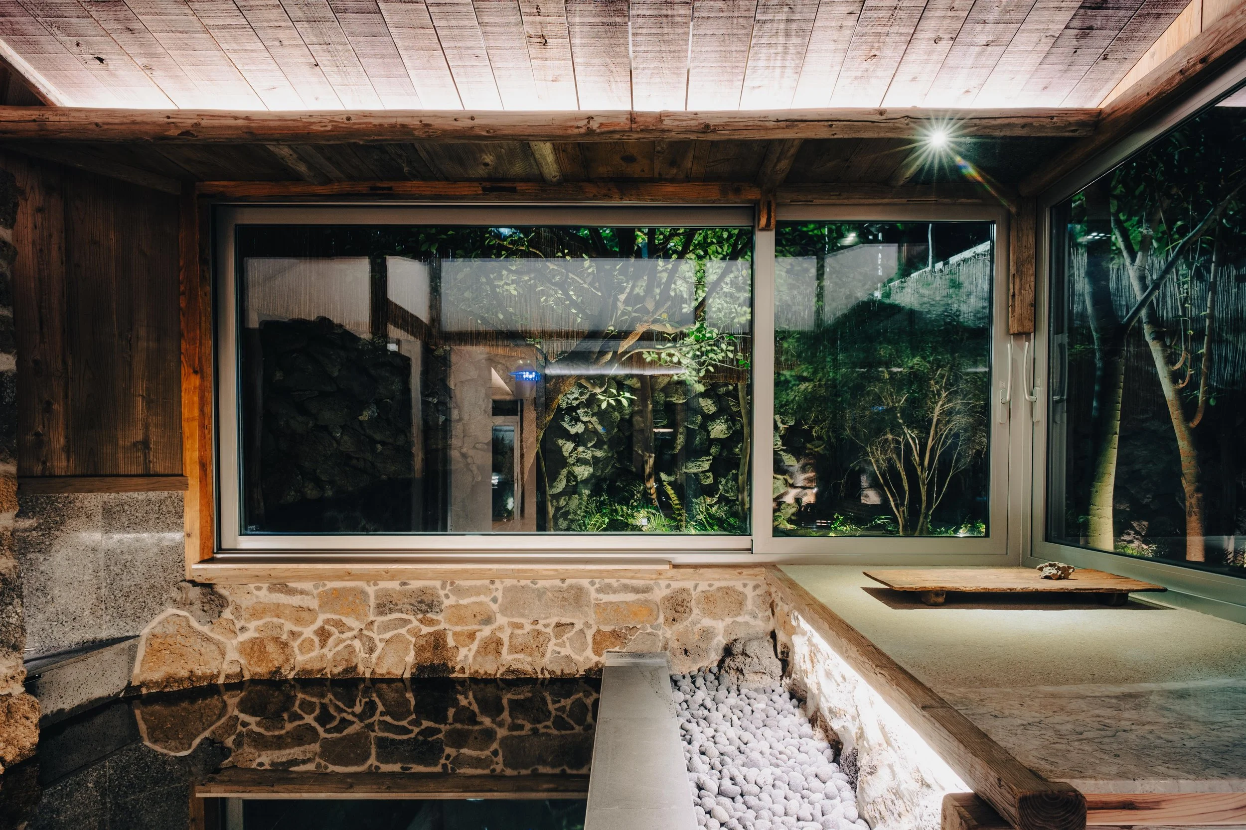 NUUT Aewol 눗애월Interior view of a room with large glass windows overlooking a garden with trees and foliage, featuring a stone wall, wooden ceiling, and a built-in stone or concrete bench with a wooden surface.