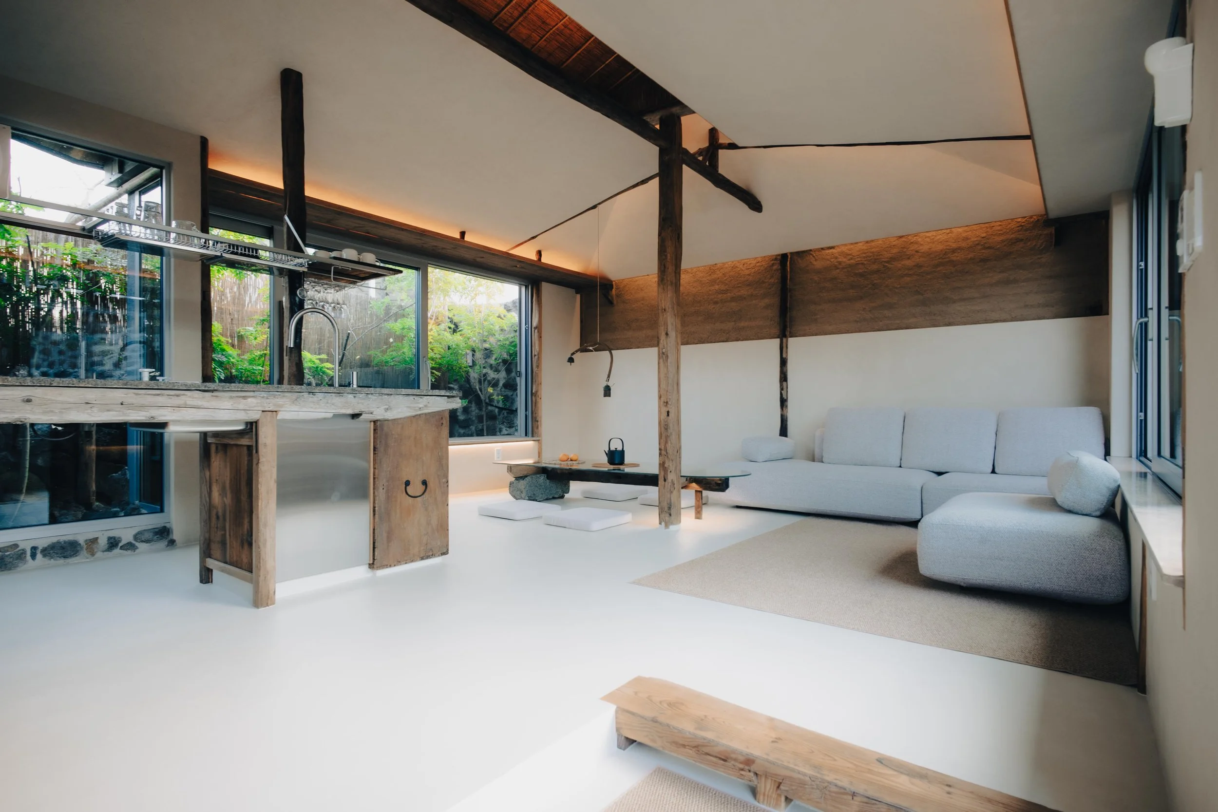 NUUT Aewol 눗애월Open-concept living space with a kitchen area featuring a rustic wooden counter, large sliding glass doors revealing greenery outside, a low white sofa with cushions, a black table with seating cushions, and wooden beams on the ceiling.