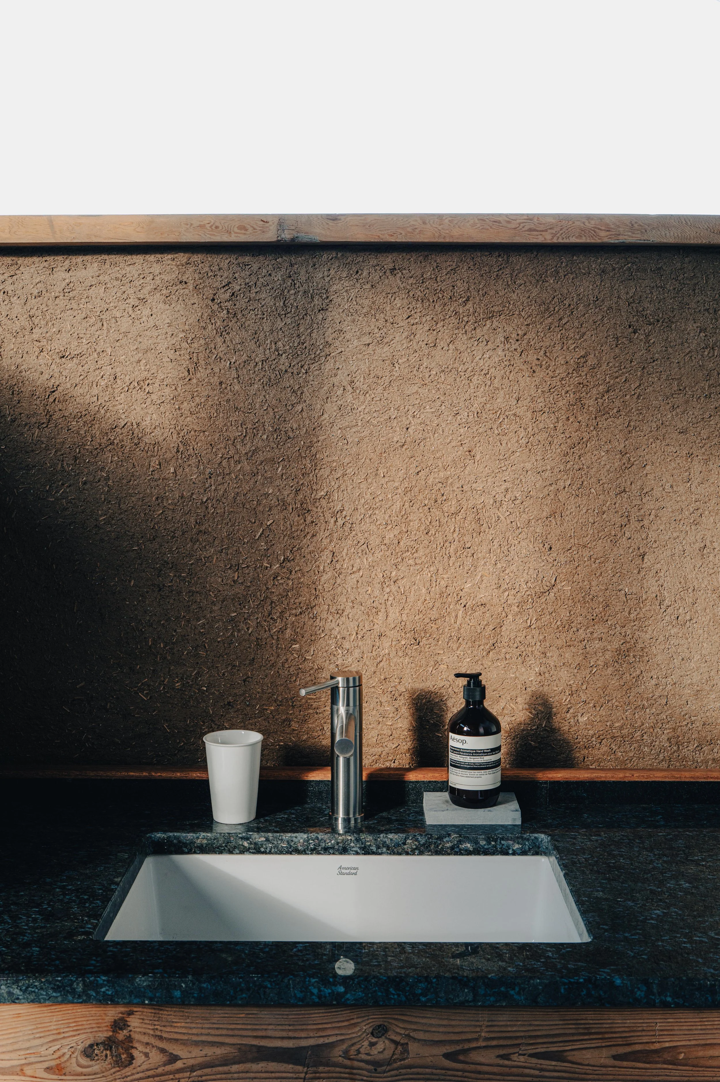 NUUT Aewol 눗애월 Bathroom sink with a white cup, a silver faucet, and a black soap dispenser on a granite countertop against a textured beige wall.