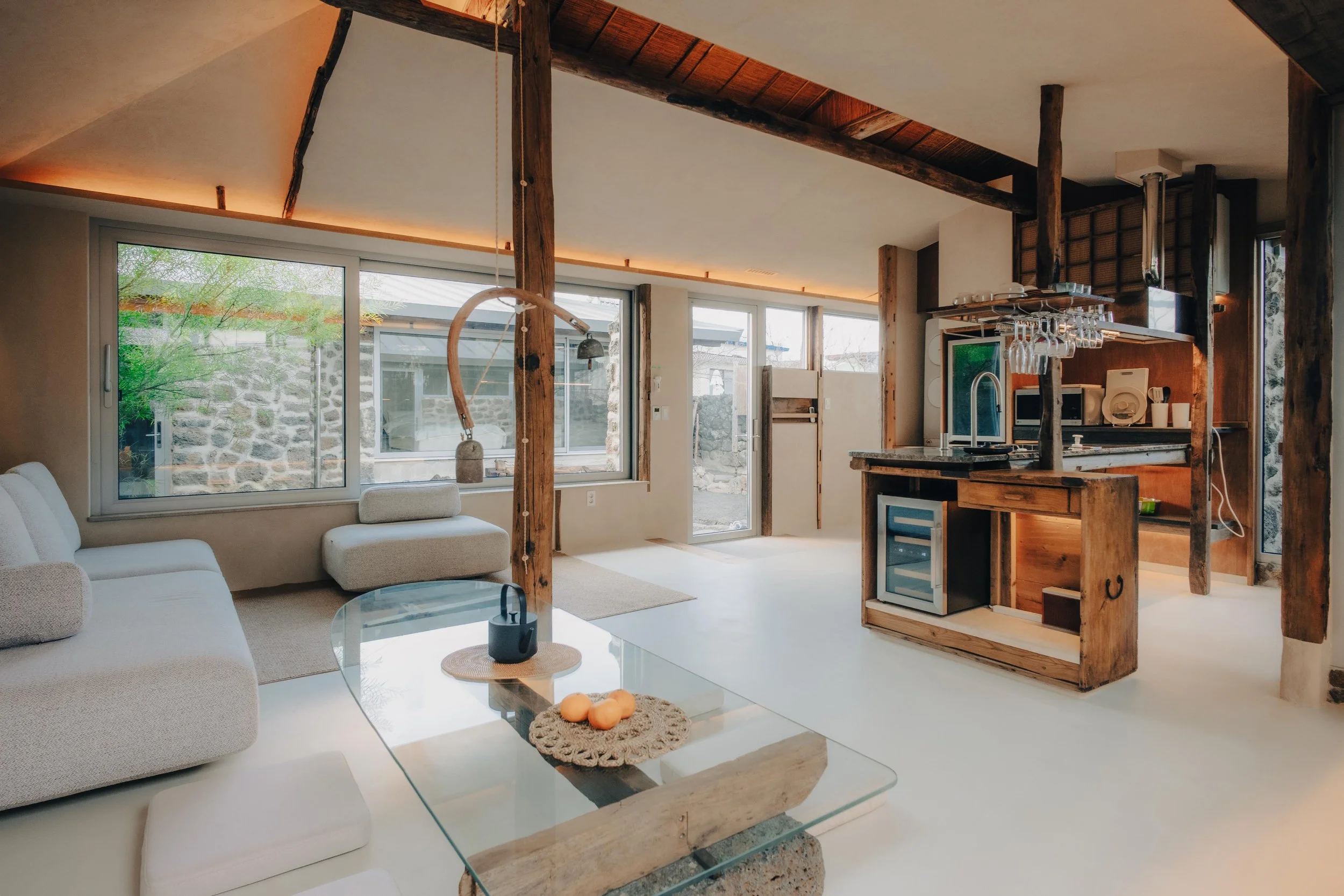 NUUT Aewol 눗애월 Open-concept living space with large windows, white sofa, glass coffee table, and wooden accents, including a kitchen area with hanging glasses and small appliances.