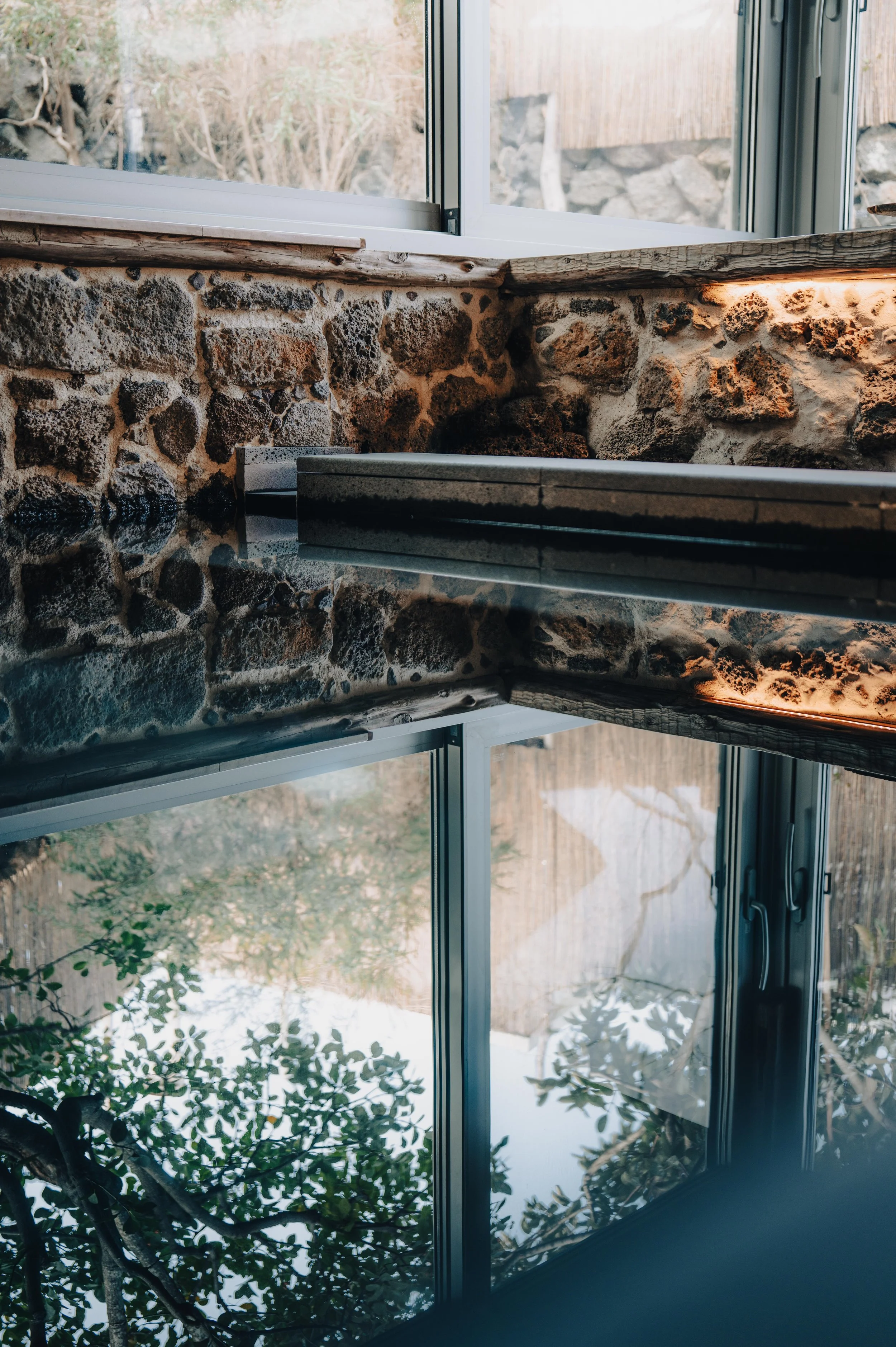 A stone wall with a window above it, reflecting trees and sky outside. The reflection in the window is visible and a small section of a black object is seen on the right side of the image.