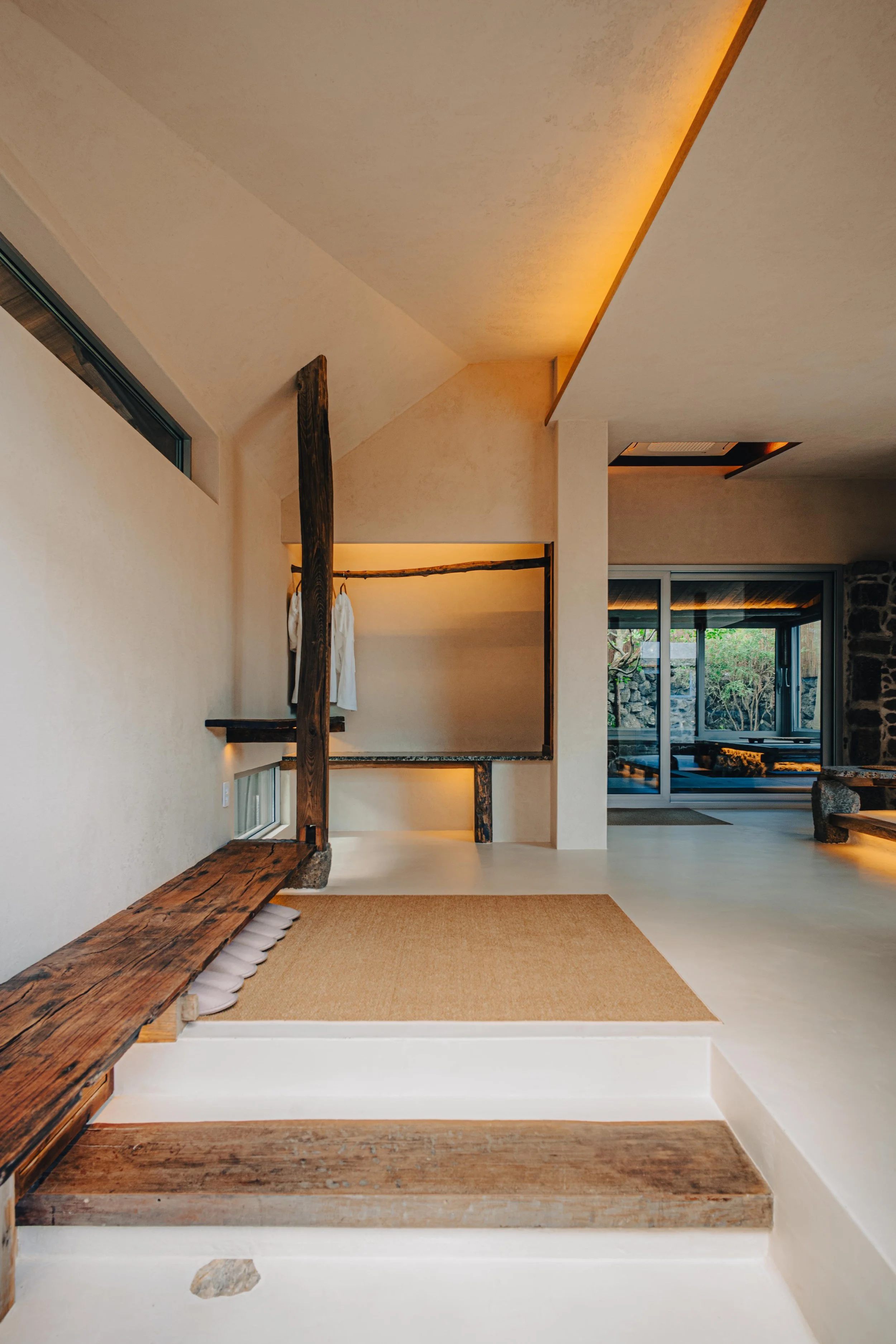 NUUT Aewol 눗애월 Interior of a modern minimalistic living space with wooden accents, a seating area with white cushions, and large glass sliding doors opening to an outdoor patio.