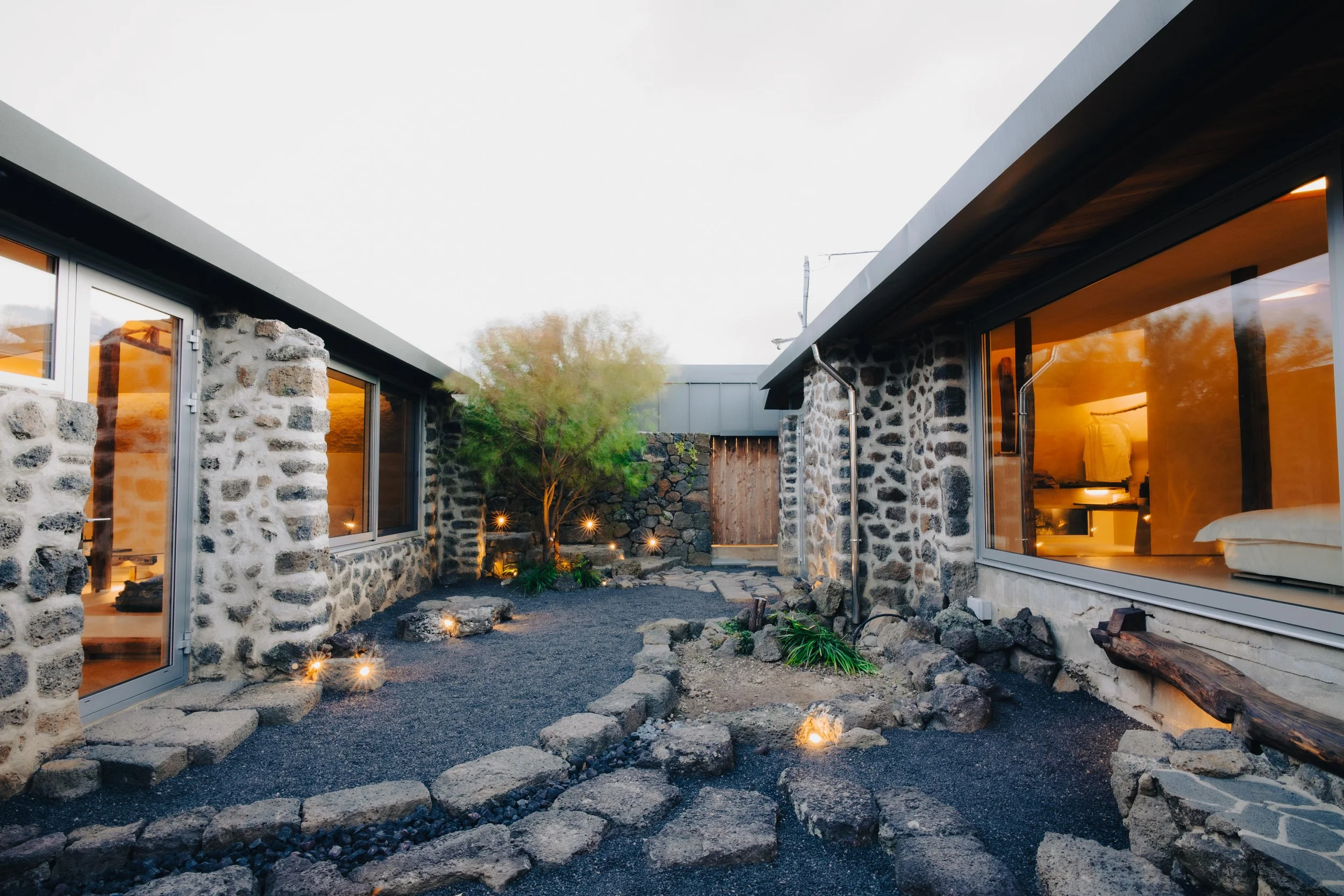 NUUT Aewol 눗애월 Modern house with stone walls, large windows, a small landscaped garden, and outdoor lighting at dusk.