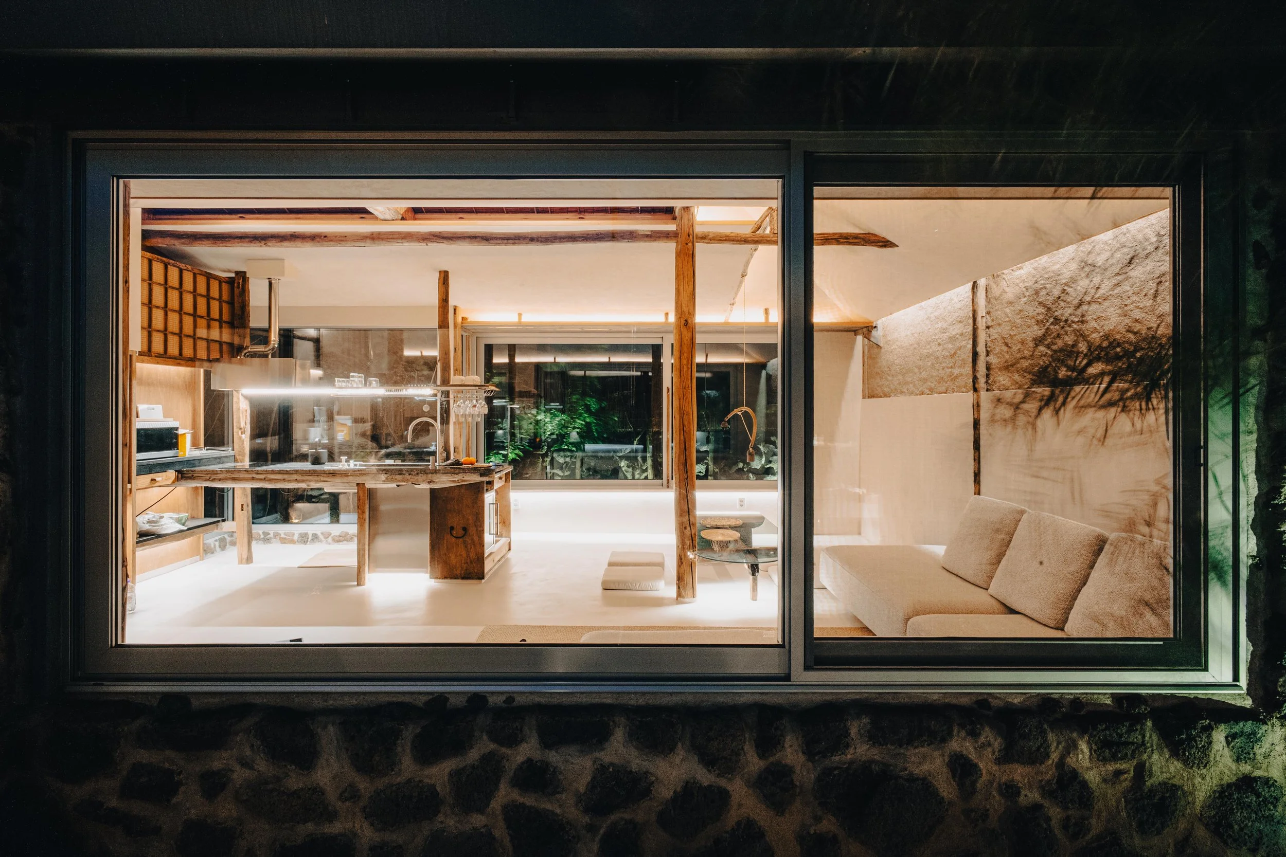 NUUT Aewol 눗애월 Interior view of a modern, minimalist living room and kitchen seen through a large window at night.