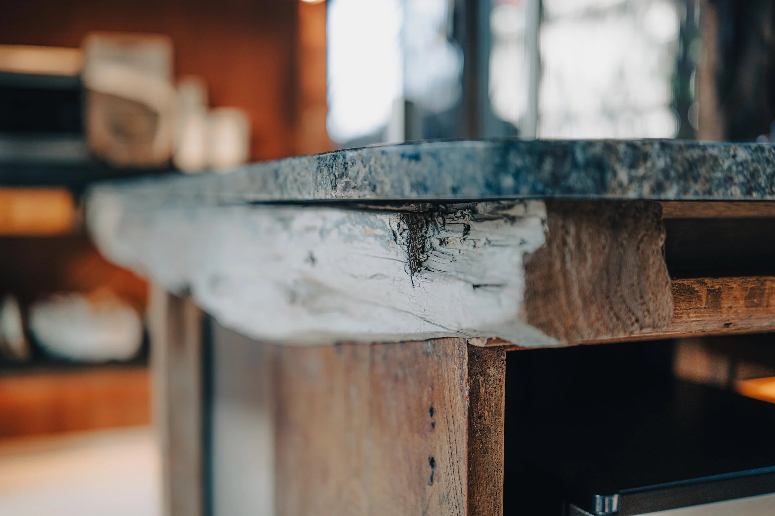 NUUT Aewol 눗애월 Close-up of a kitchen counter edge, showing a dark granite top, a white distressed wood beam support, and a wooden cabinet.