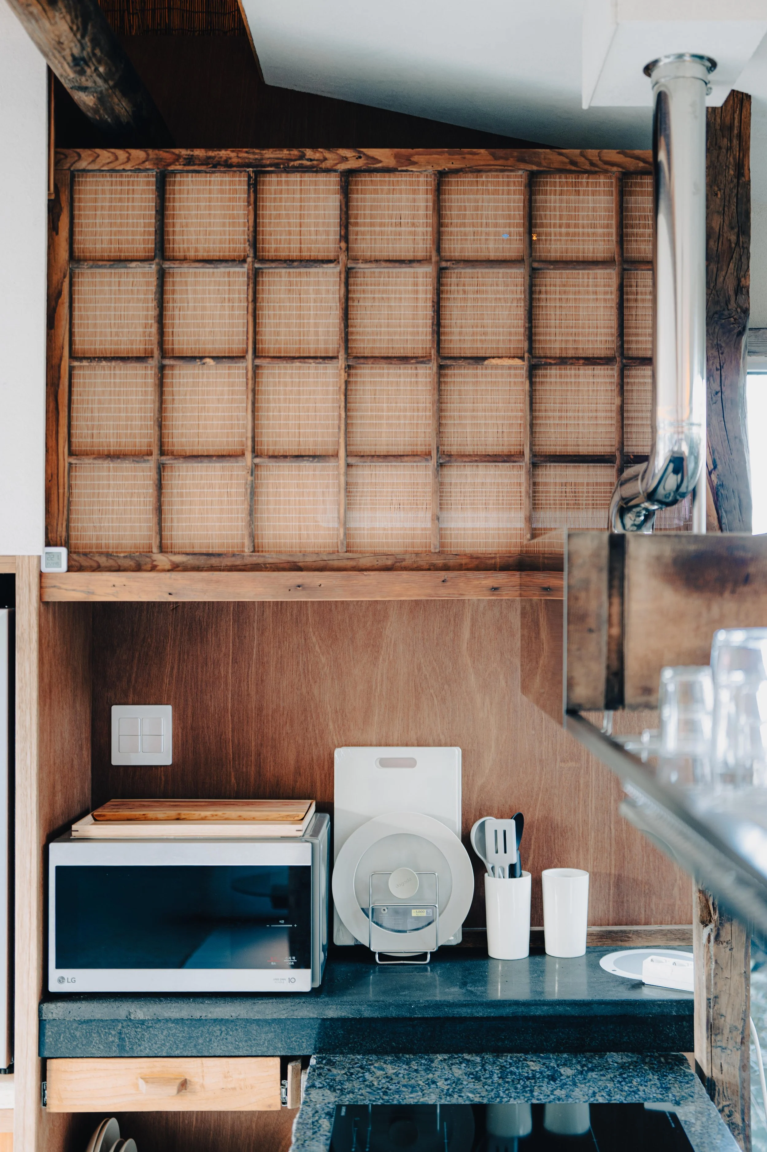 NUUT Aewol 눗애월 Looking down at a kitchen counter with a microwave, a white appliance, cups, and utensils, with wooden shelves above.