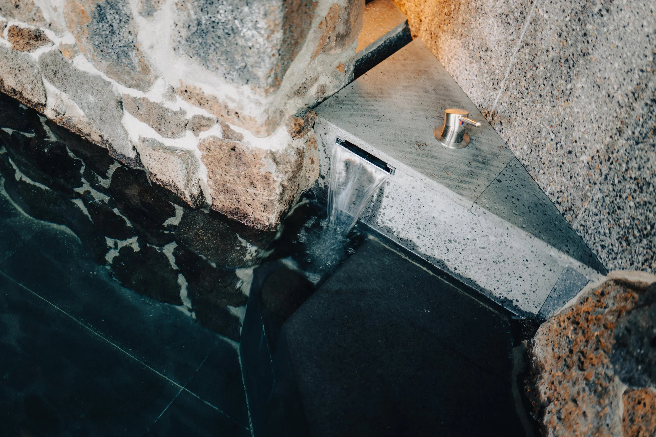 Indoor water feature with a small waterfall flowing from a slit in a stone wall into a black tiled pool, with a concrete countertop and a modern faucet nearby.