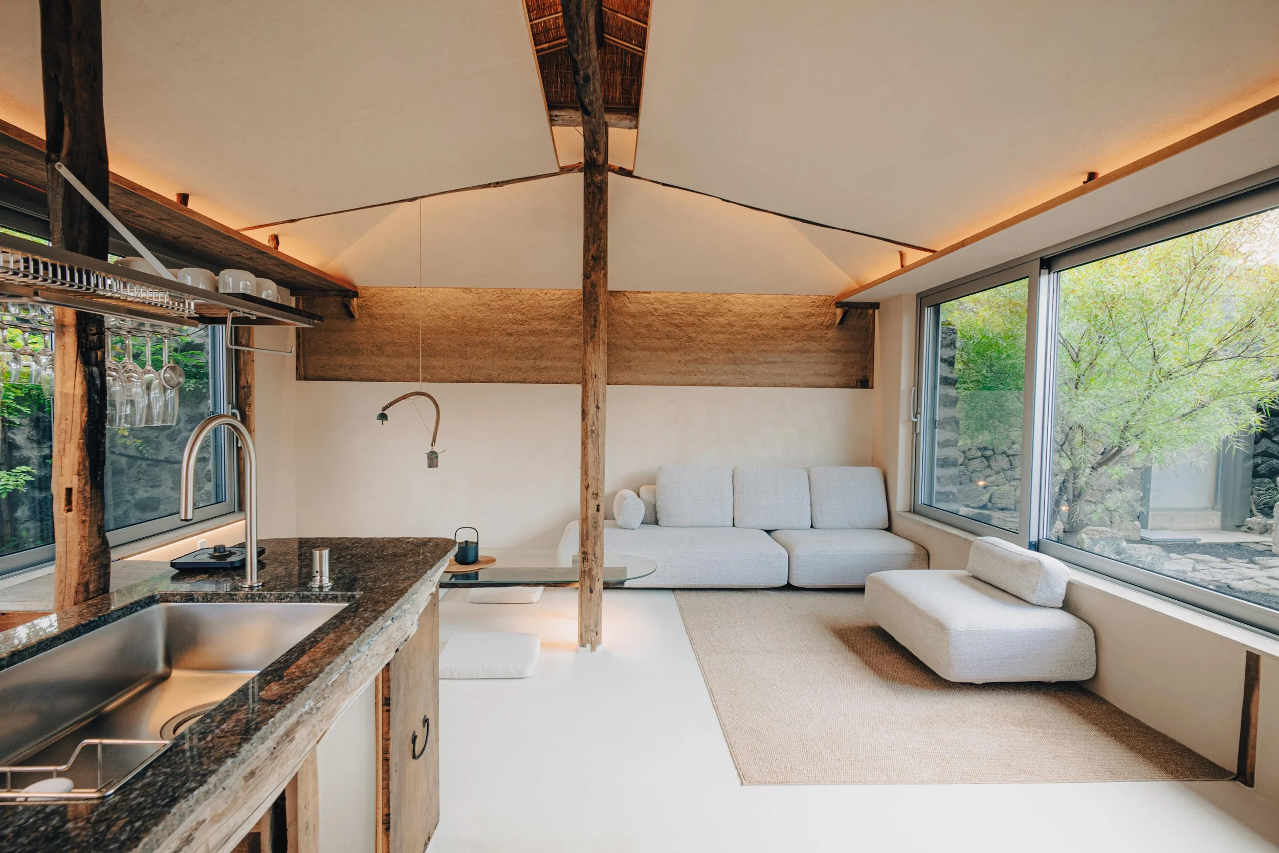 NUUT Aewol 눗애월A cozy living room with large windows, a light grey sofa, and a small ottoman. The room has exposed wooden beams and a neutral color palette. The kitchen area is partially visible with a granite countertop and hanging cups.