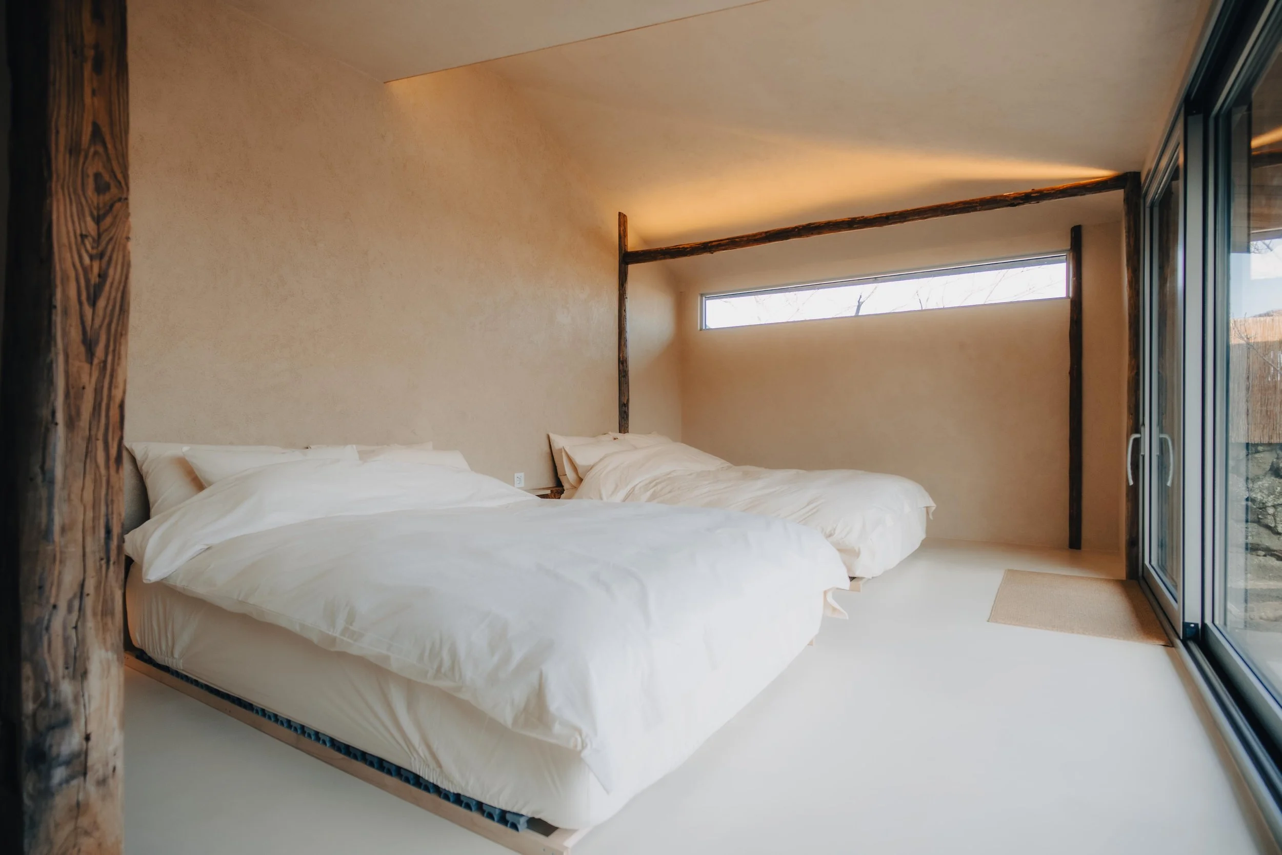 NUUT Aewol 눗애월 A minimalist bedroom with two unmade beds and white bedding, beige walls, horizontal window, and glass sliding door leading outside.