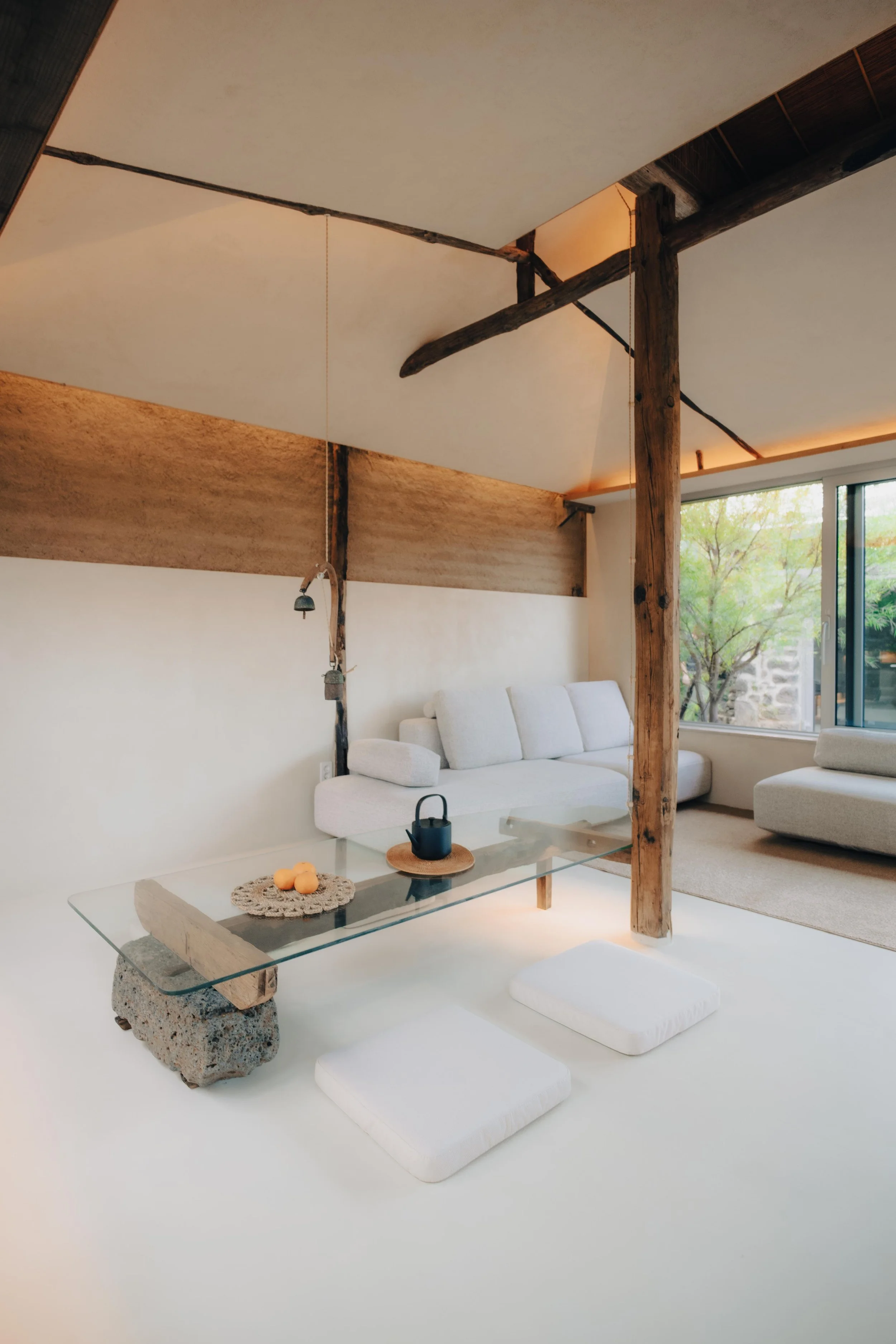 NUUT Aewol 눗애월 Minimalist living room with white sofas, a glass coffee table on a white floor, and wooden beams supporting the ceiling. Large window shows greenery outside.