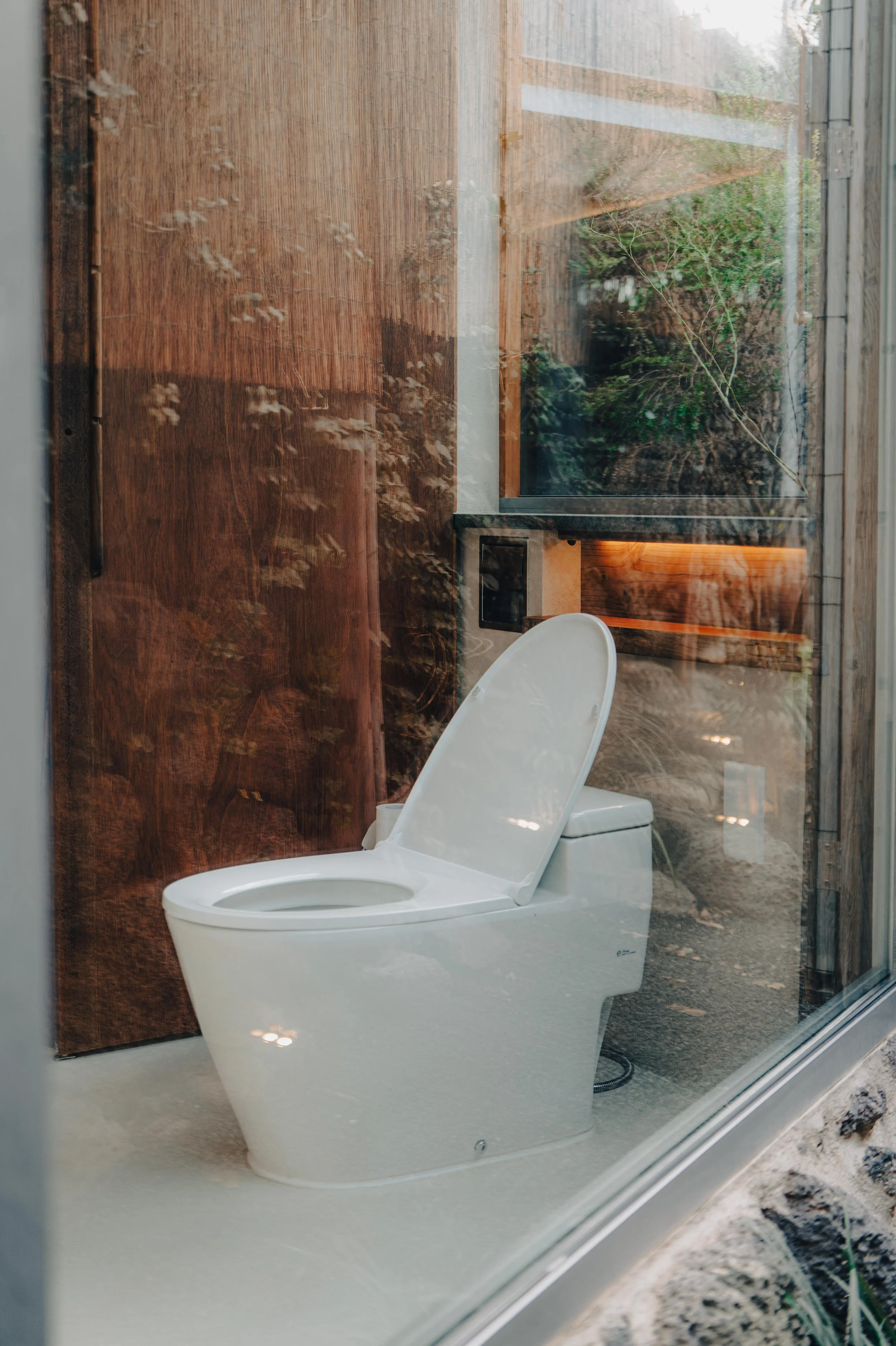 NUUT Aewol 눗애월 Toilet inside a room with a glass wall, with a wood wall and greenery visible outside.