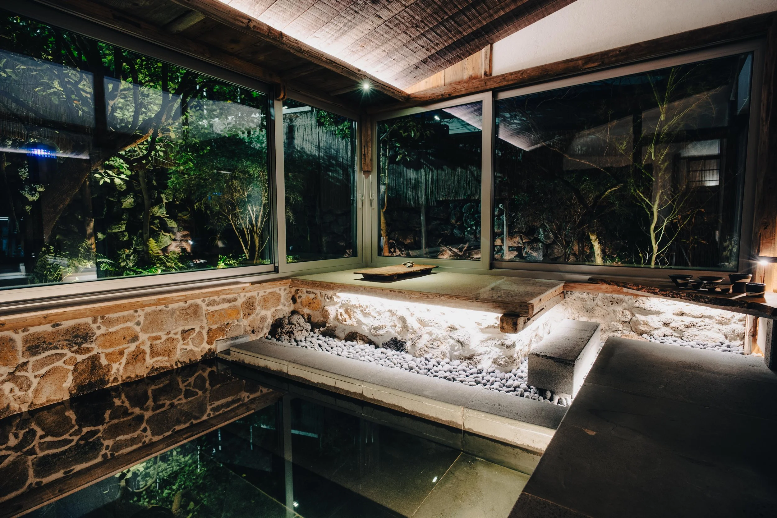 NUUT Aewol 눗애월 Interior of a room with floor-to-ceiling windows showing a garden outside, featuring trees and rocks, with a stone wall, wooden ceiling, and a raised table with stones and pebbles underneath.
