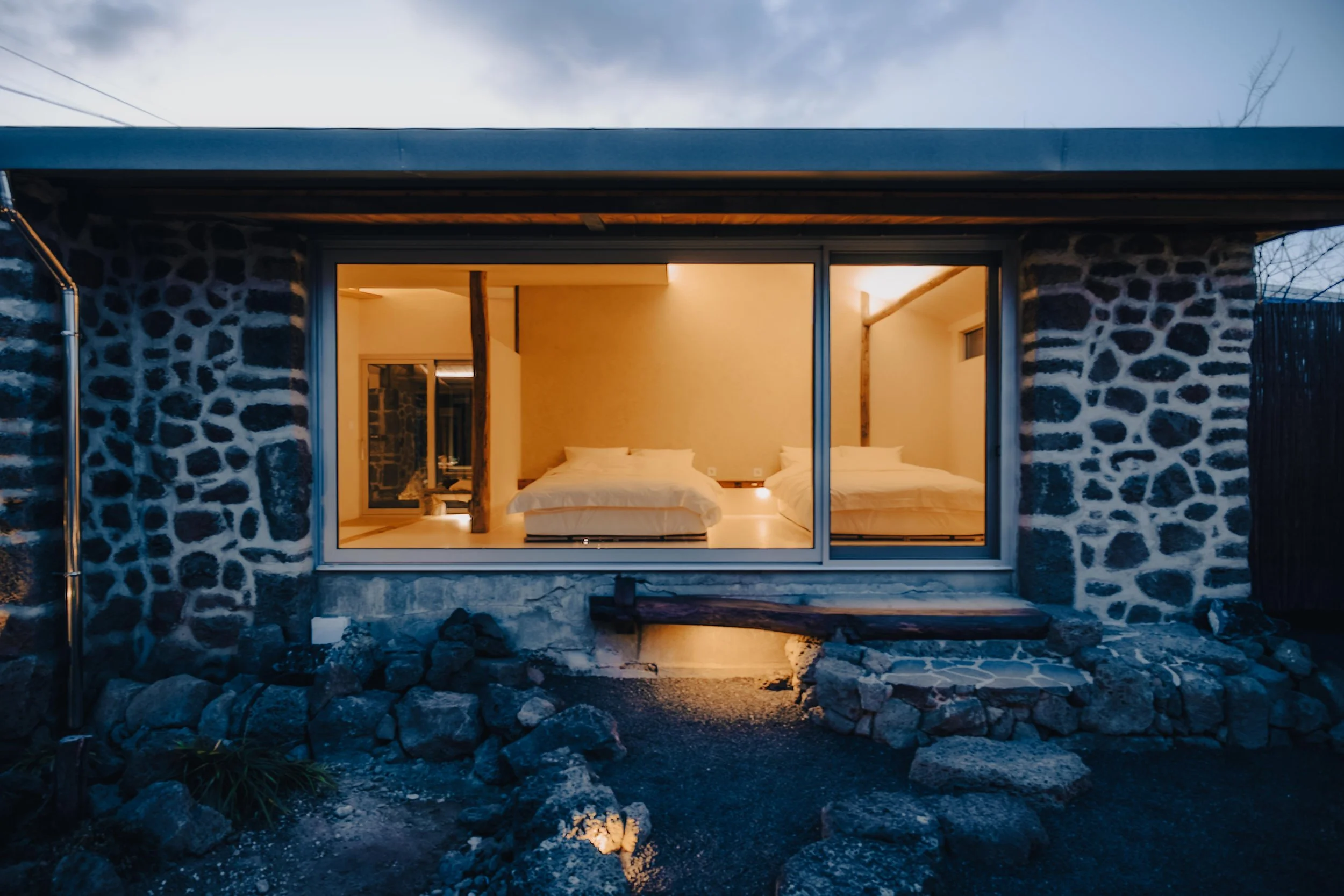 NUUT Aewol 눗애월 Exterior view of a modern stone building at night with illuminated interior showing two beds through large window