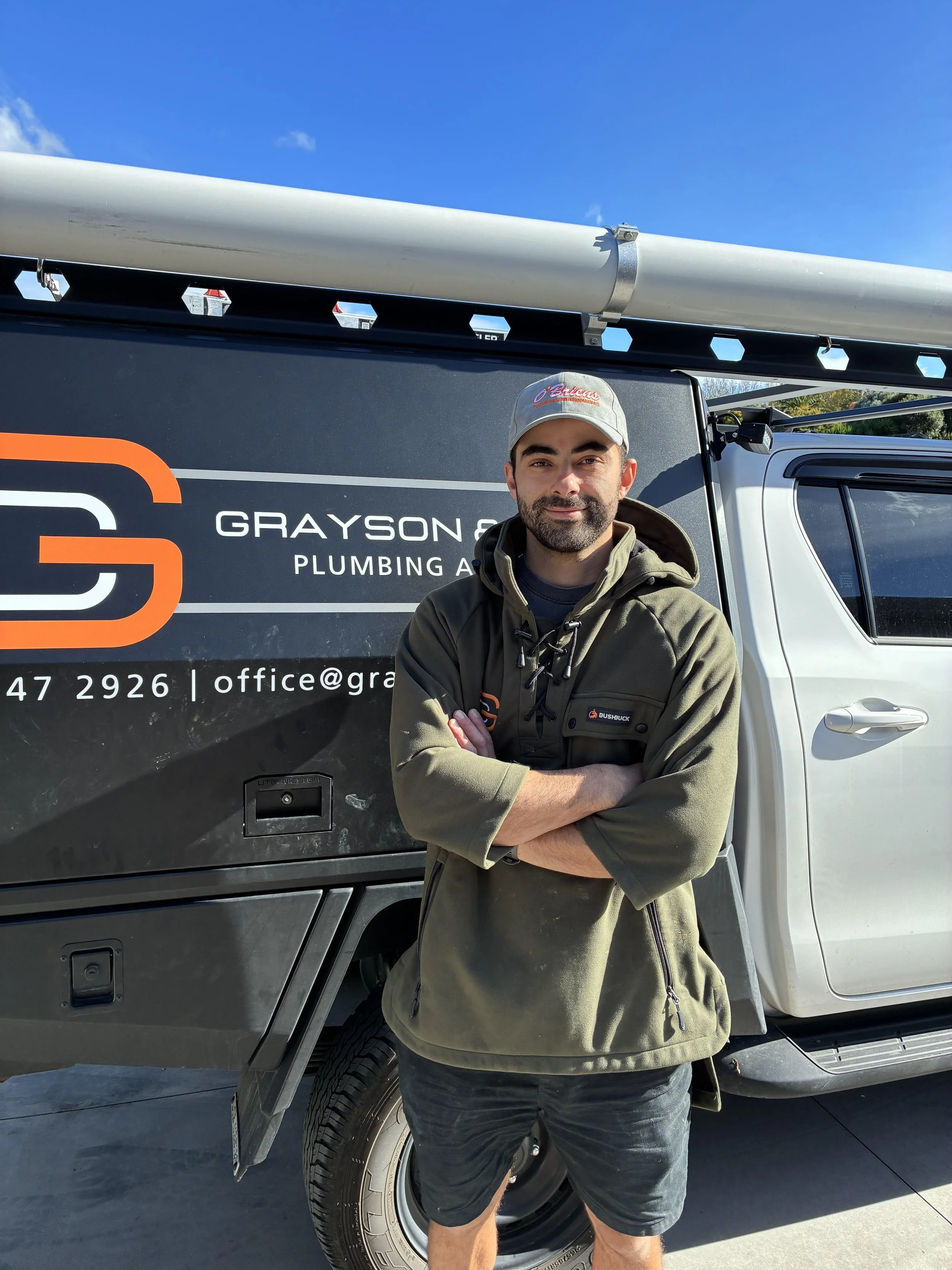 A man posing with his arms crossed in front of a service van with the logo 'Grayson & Plumbing' and a large pipe on top.