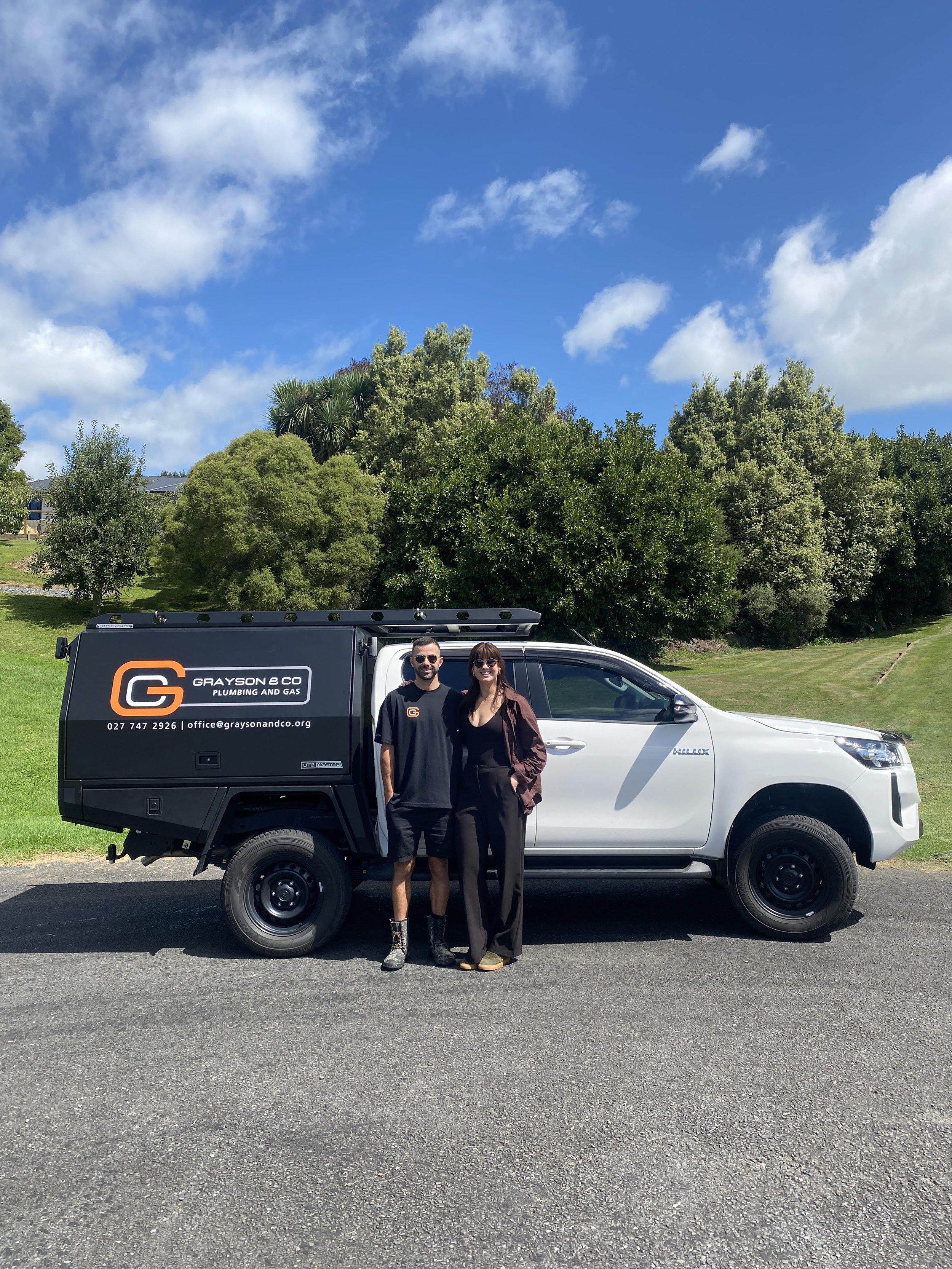 A man and woman standing in front of a white utility truck with a black service body, on a paved road with grass and trees in the background, under a blue sky with scattered clouds.