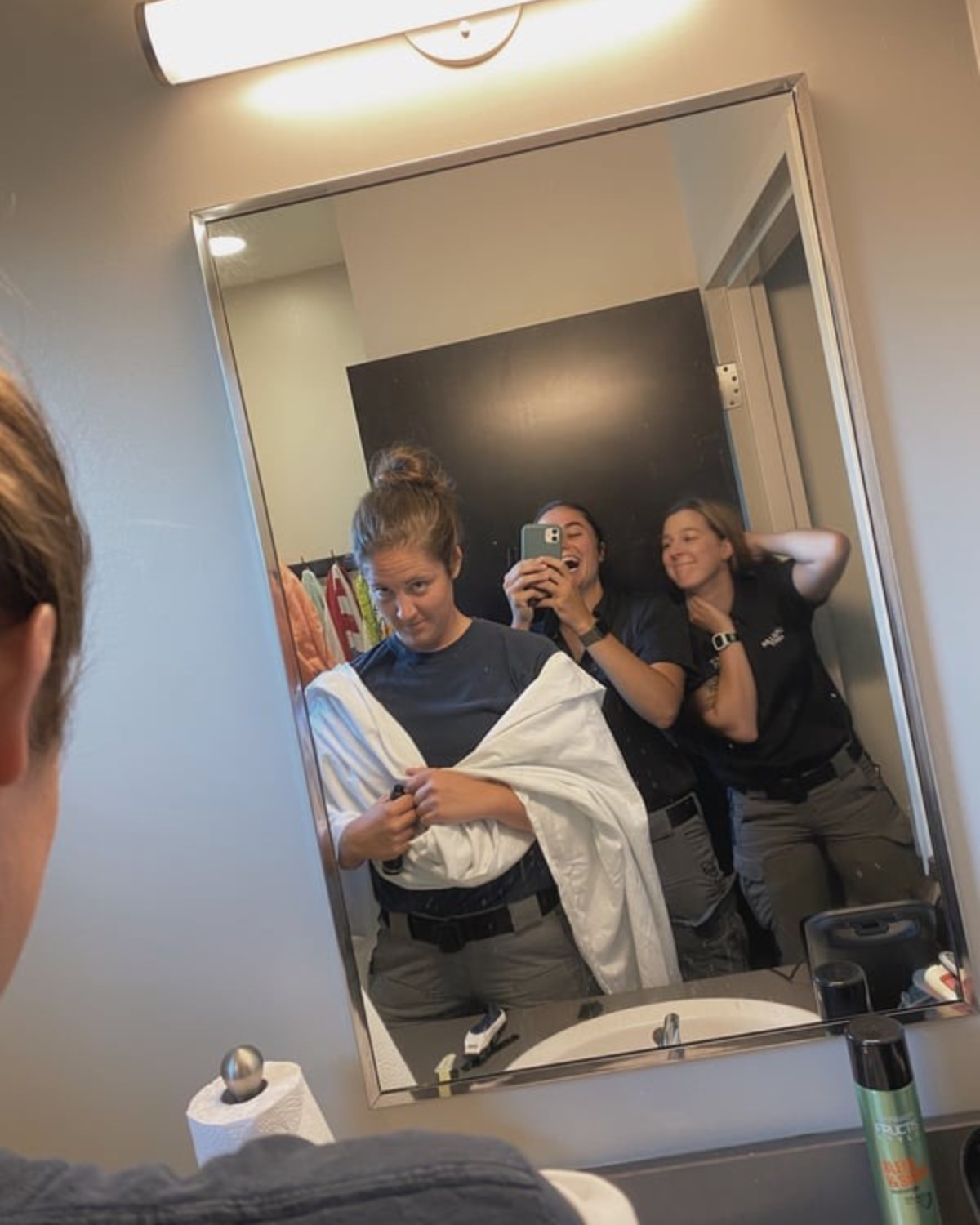 Three young women taking a mirror selfie in a bathroom, with one holding a blanket and making a funny face, while the others are smiling and laughing.