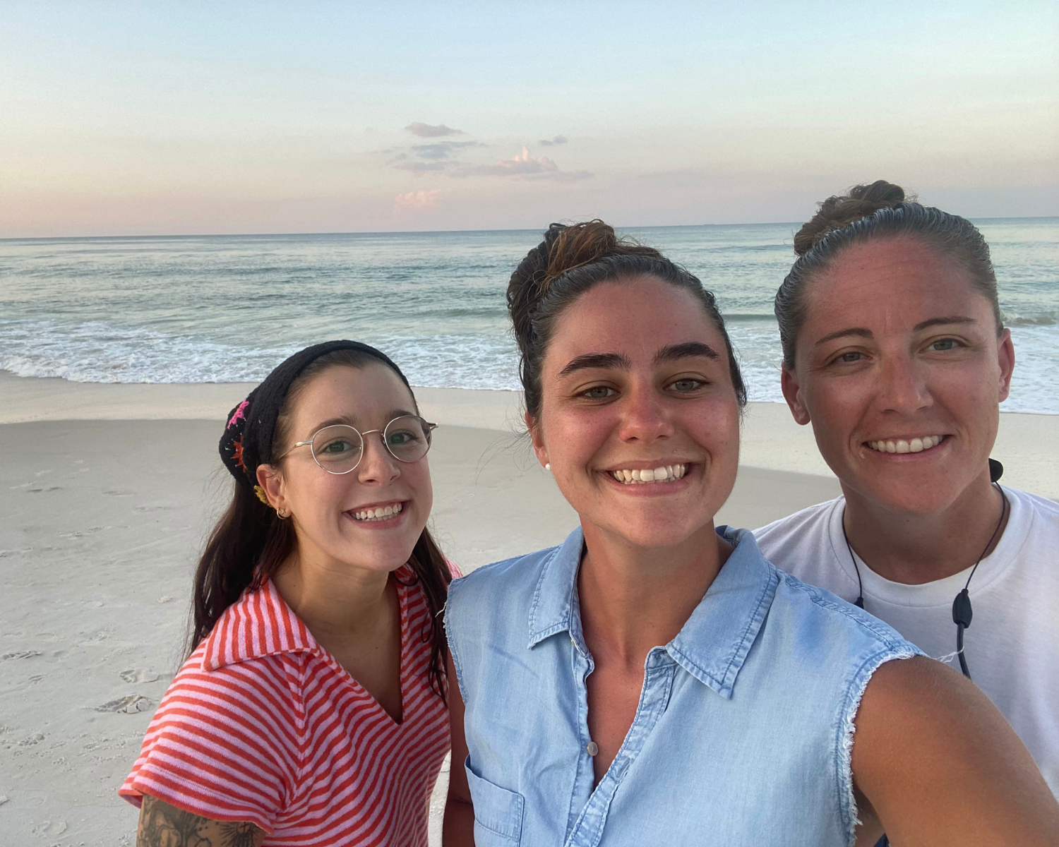 Three women smiling and taking a selfie on a beach with the ocean and a pastel-colored sky in the background