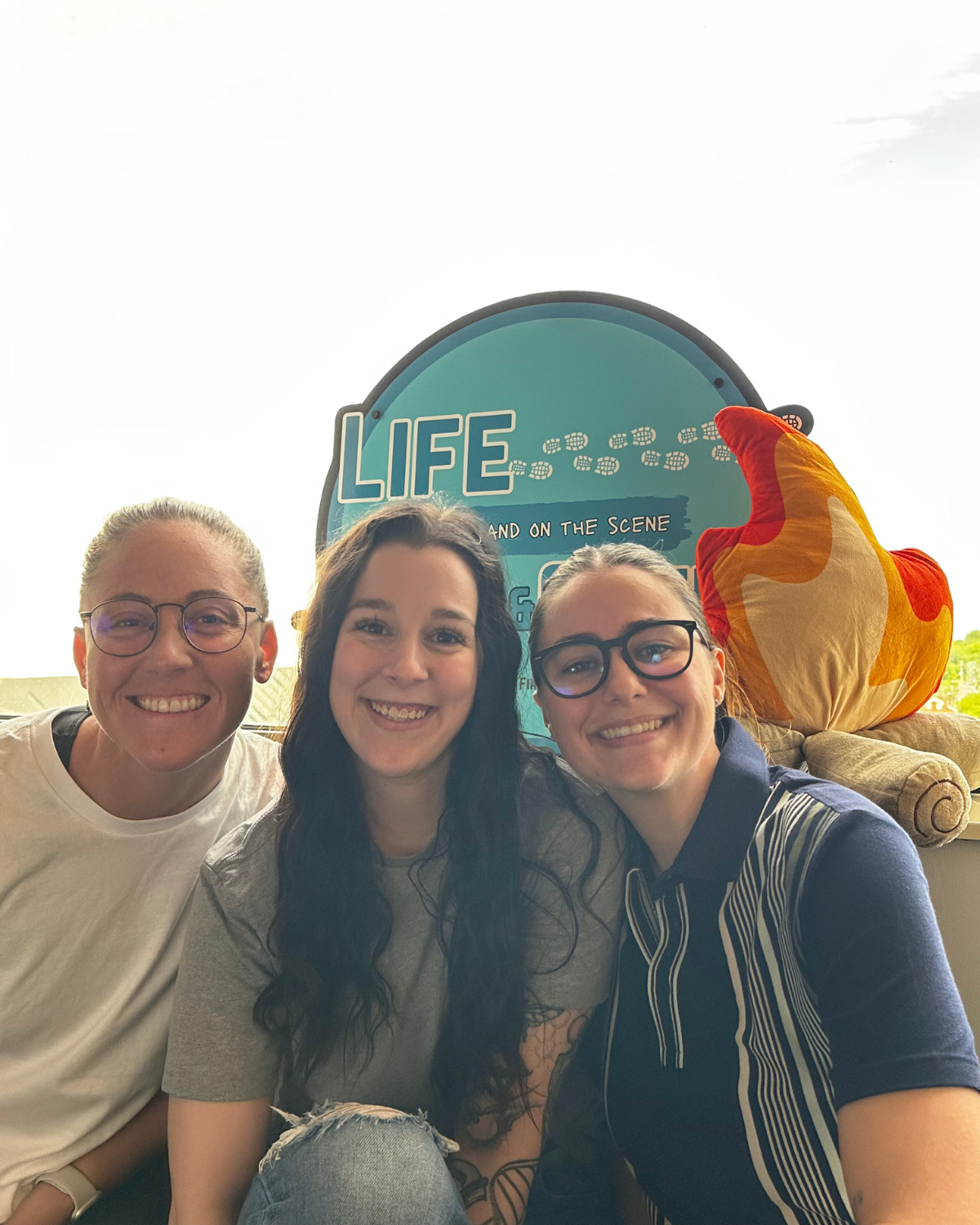 Three smiling women sitting together on a bench with a bright sky behind them, in front of a blue sign that reads 'LIFE'. There is a plush flame pillow on the right.