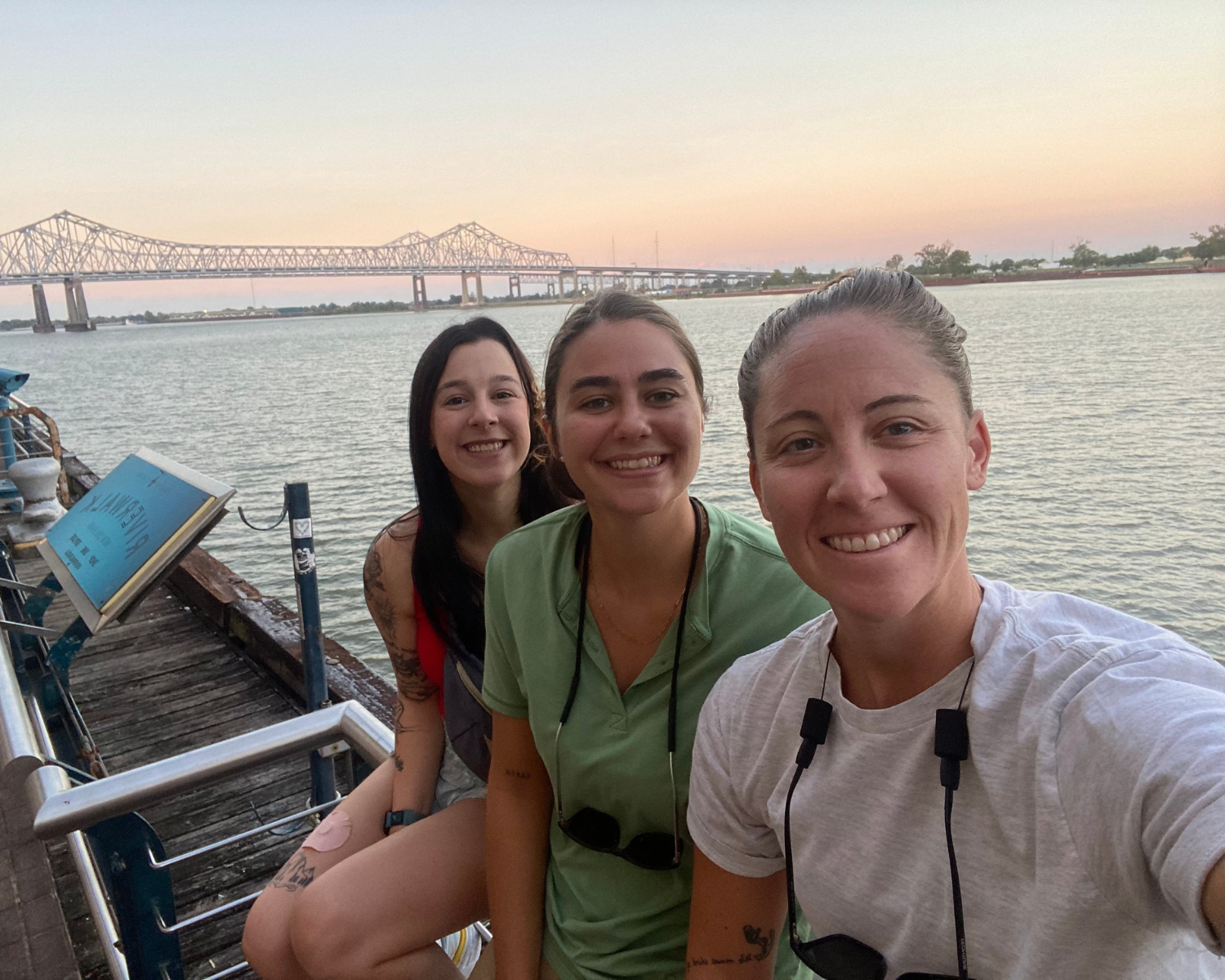 Three women smiling for a selfie on a dock beside a river, with a bridge in the background during sunset.