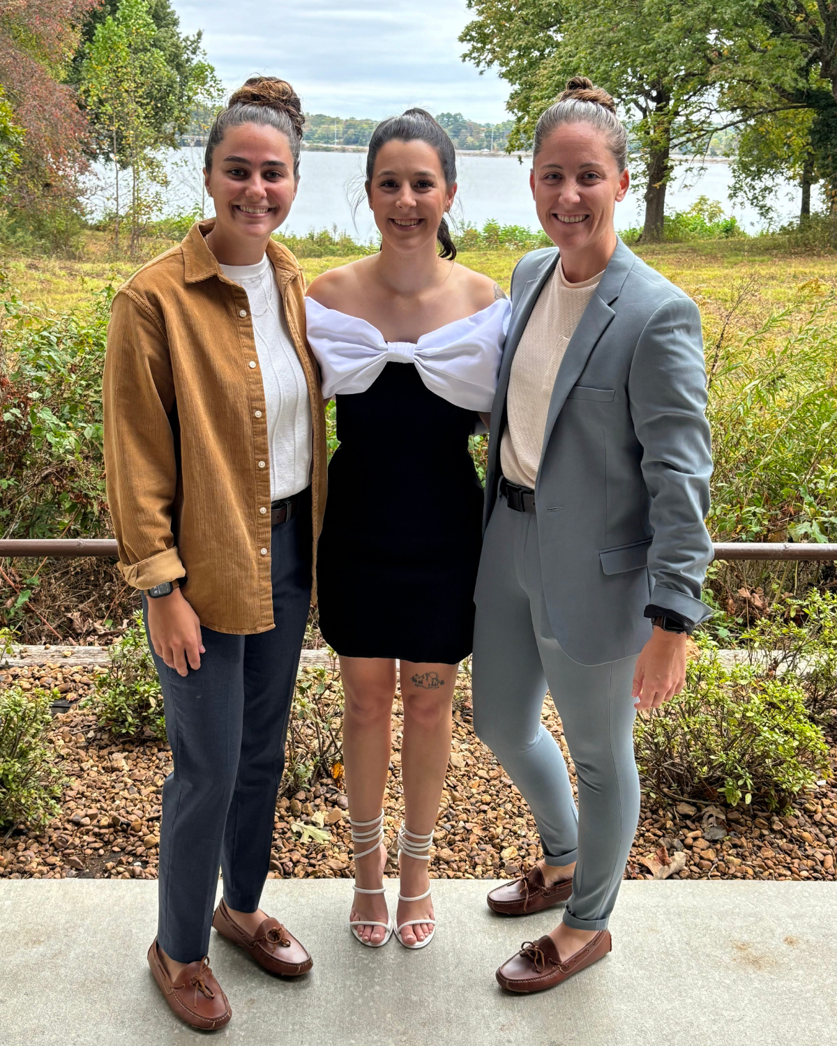 Three women standing outdoors near a body of water with trees in the background, smiling at the camera.