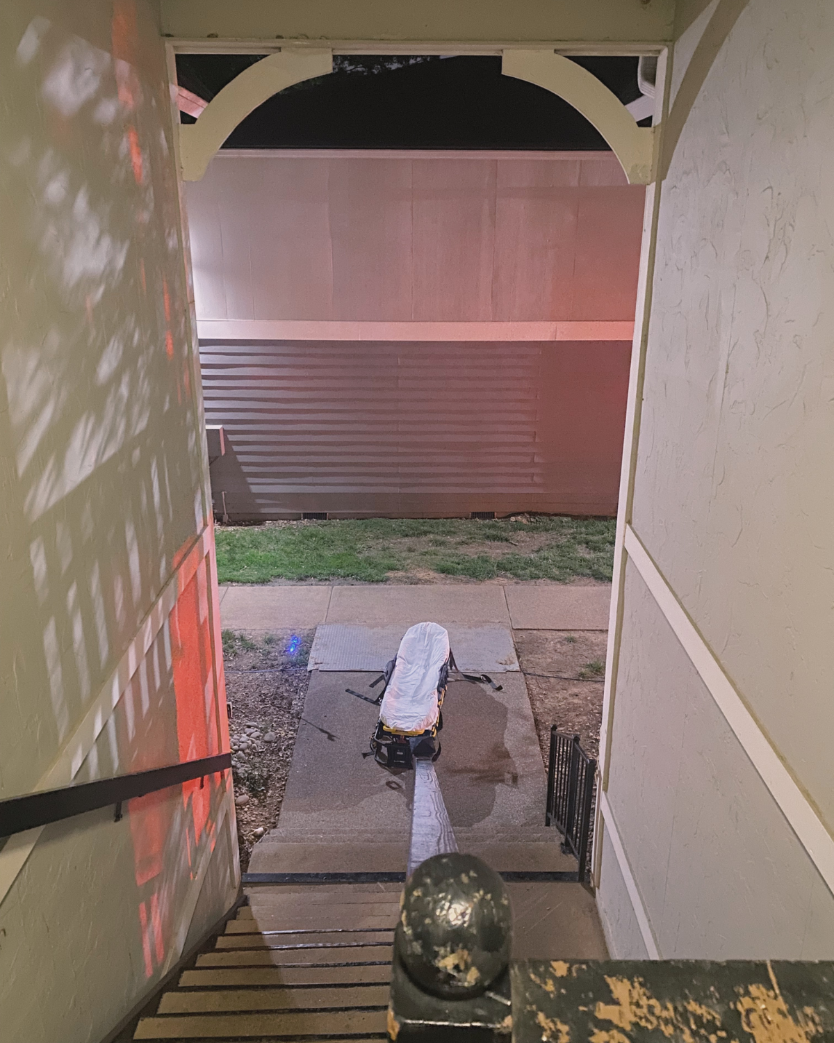 View from the top of a staircase looking outside at a sidewalk, a covered stretcher, and a closed gate.