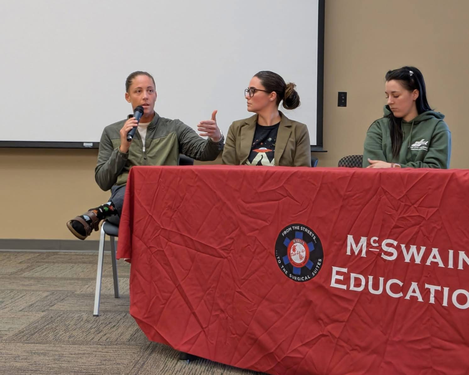 Three women sitting at a panel discussion behind a red tablecloth with 'McSwaine Education' logo, one woman speaking into a microphone and gesturing with her hand.