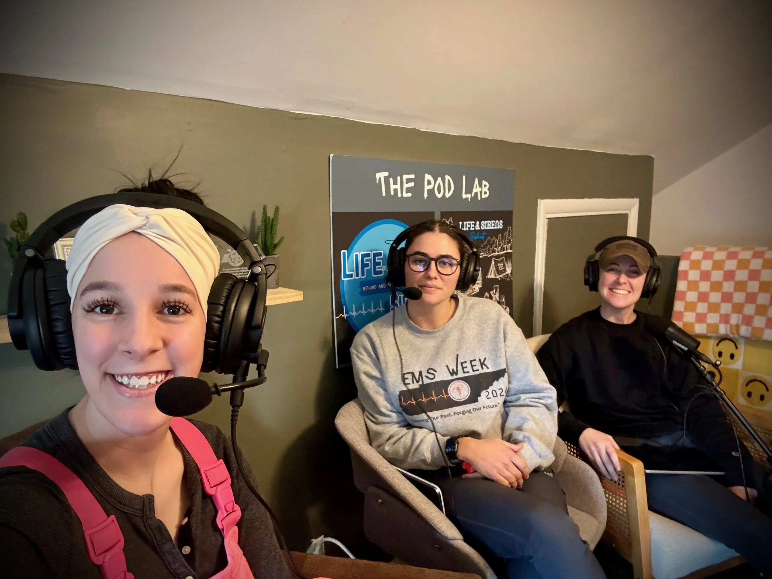 Three women wearing headphones and microphones sitting in a recording studio with a sign that reads 'The Pod Lab' behind them.