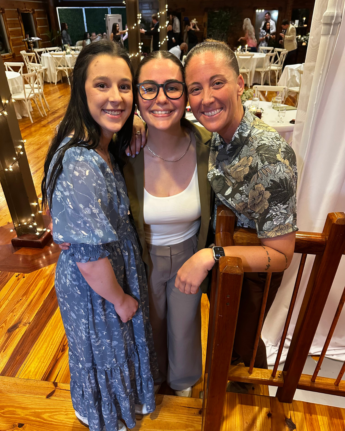 Three women smiling and posing together at a social event or celebration in a decorated indoor venue with tables, chairs, and string lights.