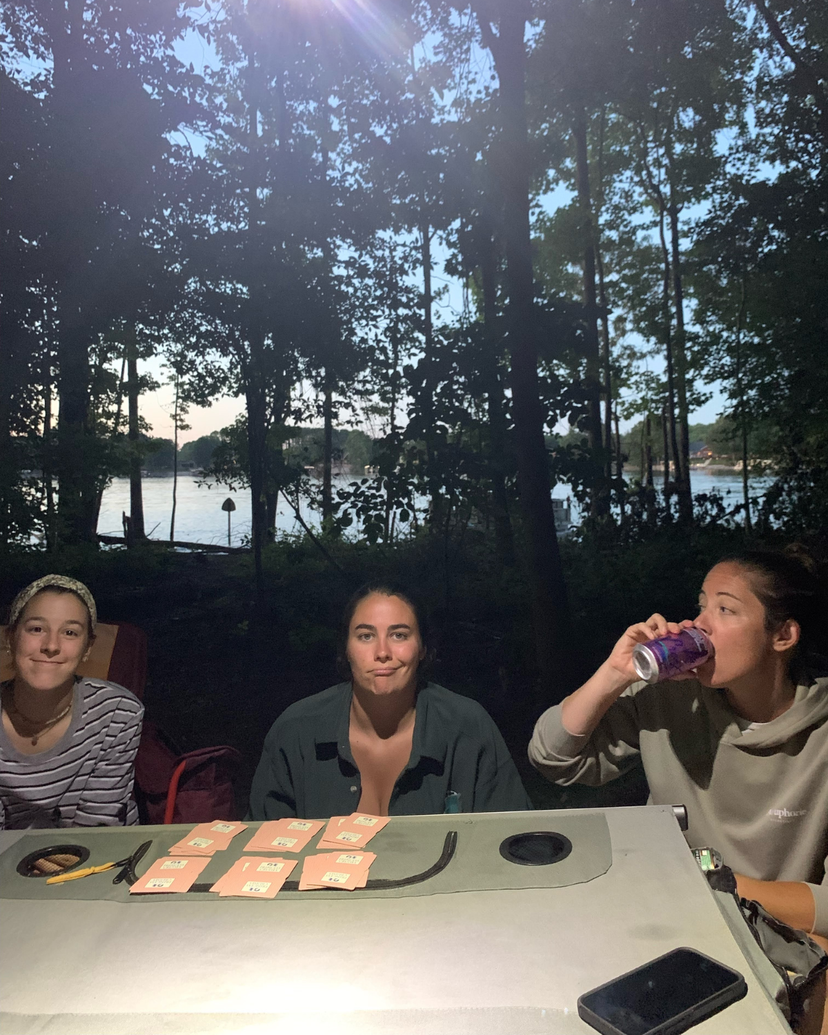 Three women sitting at a table outdoors in a wooded area near a lake during sunset. The table has cards and a pair of scissors on it. One woman is drinking from a can, while the others look at the camera.