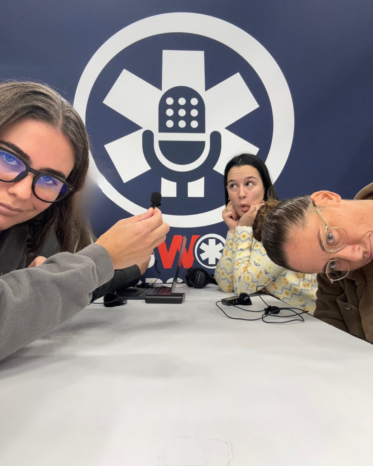 Three women at a table with microphones, posing for a selfie in front of a logo featuring a microphone and a medical symbol.