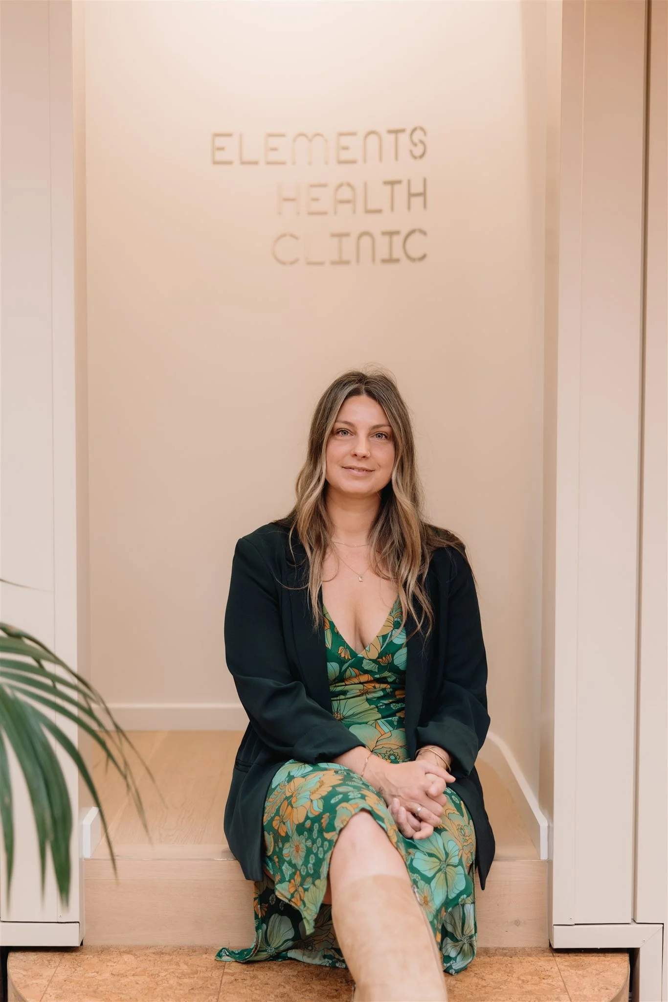 A woman sitting on a step inside Elements Health Clinic, wearing a green floral dress and black blazer, with a neutral expression on her face.