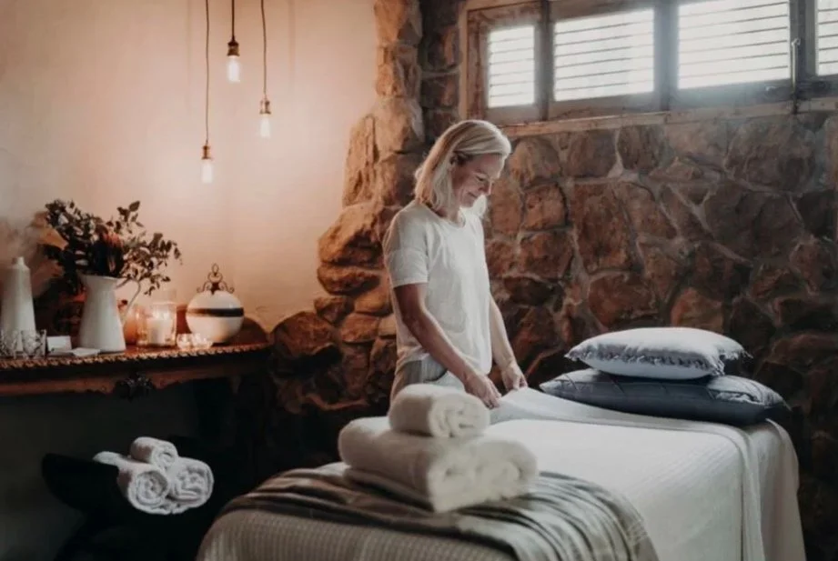 A woman in a white top making a bed in a cozy room with a stone wall, a window, and a wooden shelf with flowers, candles, and decor.