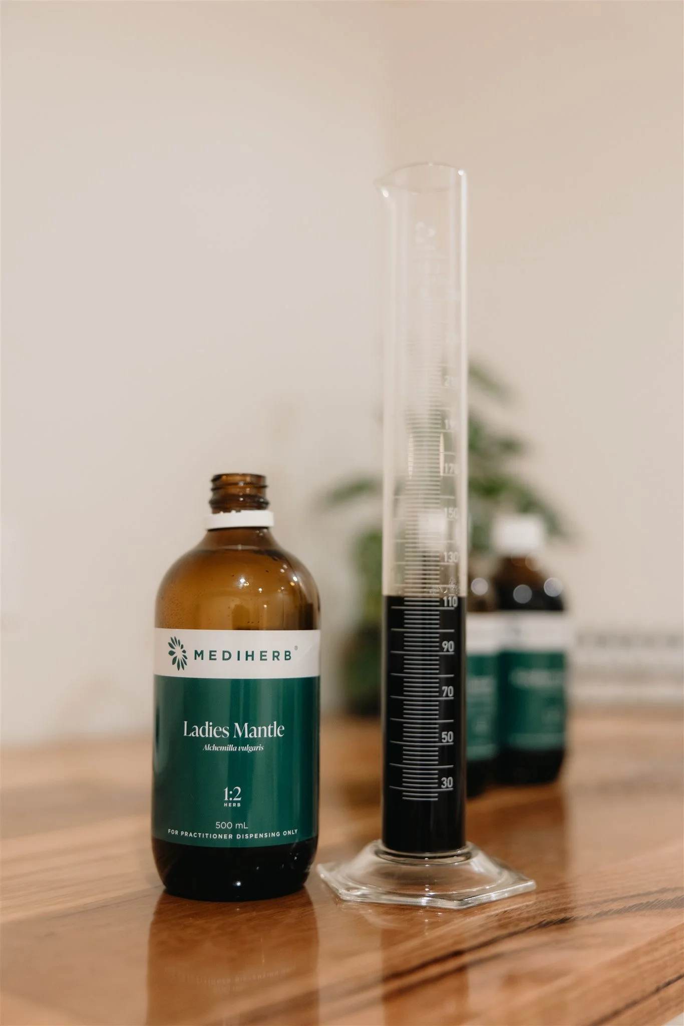 A brown bottle labeled "Mediherb Ladies Mantle" next to a large graduated measuring cylinder filled with a dark liquid, on a wooden surface.
