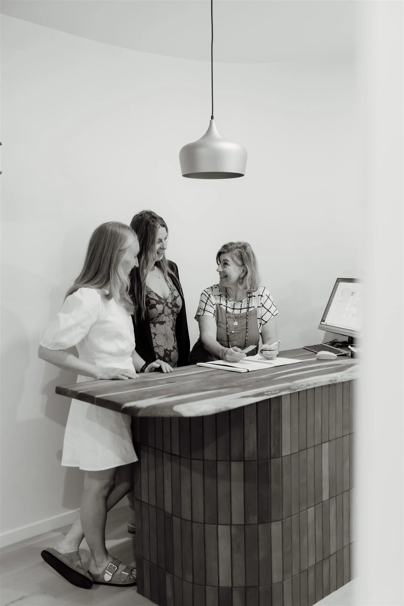 Four women standing and sitting around a wooden reception desk in a modern setting, engaged in conversation, with a computer and notepad on the desk.