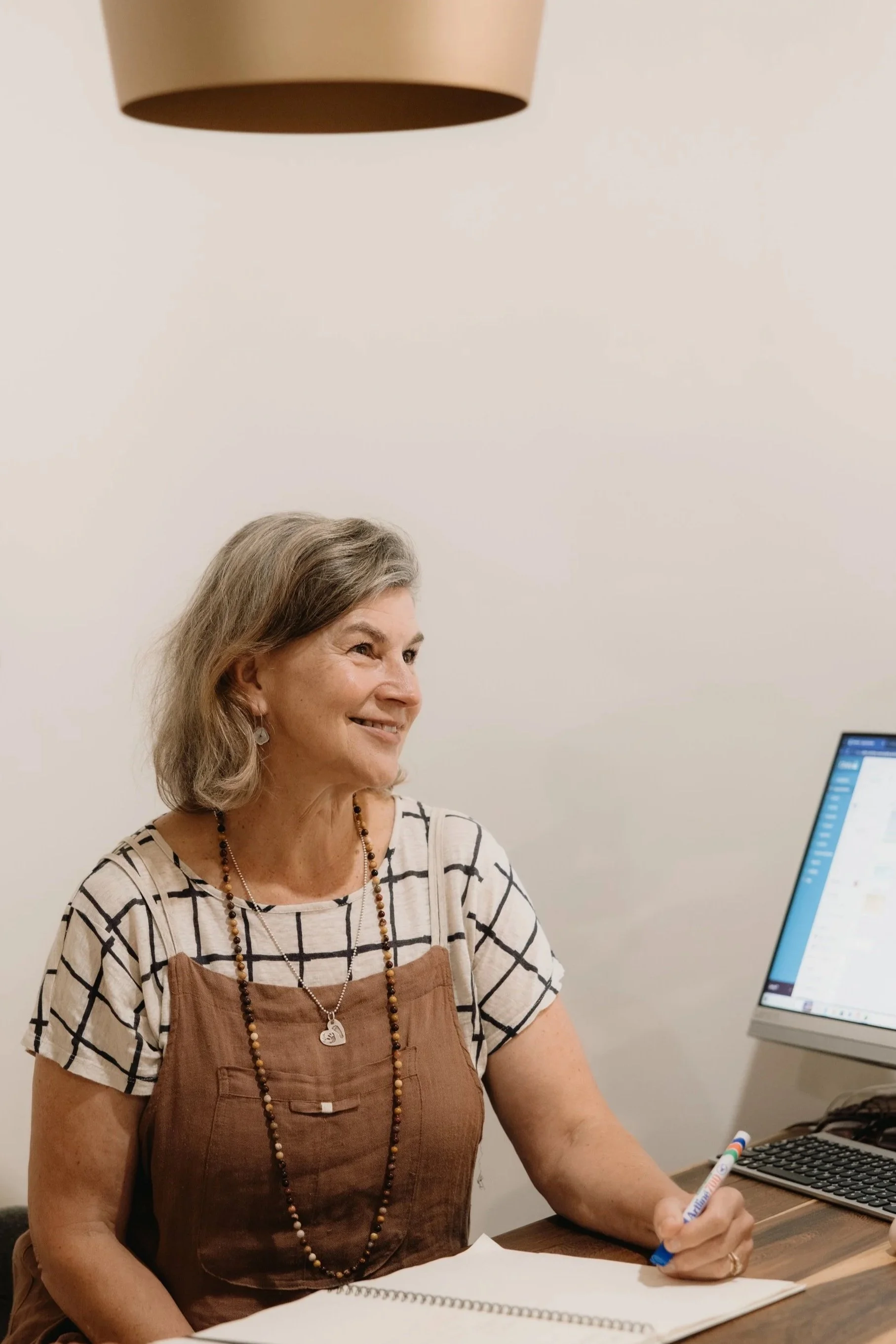 A woman with gray hair and a warm smile seated at a desk, writing on a sheet of paper with a pen. She is wearing a plaid shirt layered with a brown apron and a beaded necklace. There is a computer monitor and a keyboard to her right, and a colorful pen on the desk. The background is a plain white wall.