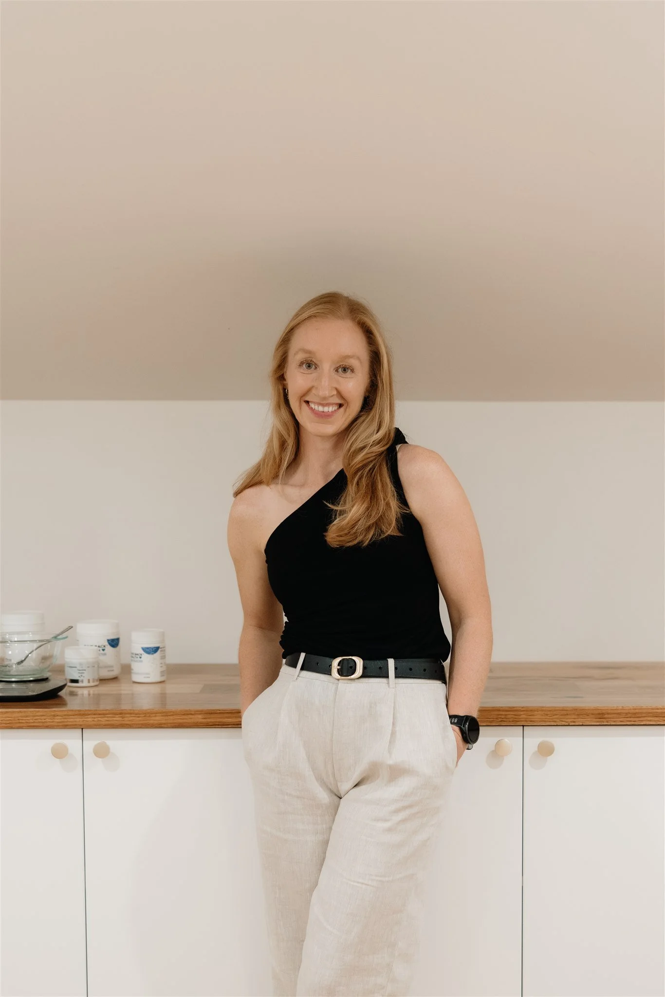 A young woman with long red hair smiling, wearing a black sleeveless top and light-colored pants, standing in a kitchen with a wooden countertop and white cabinets, with several skincare or supplement containers on the counter.