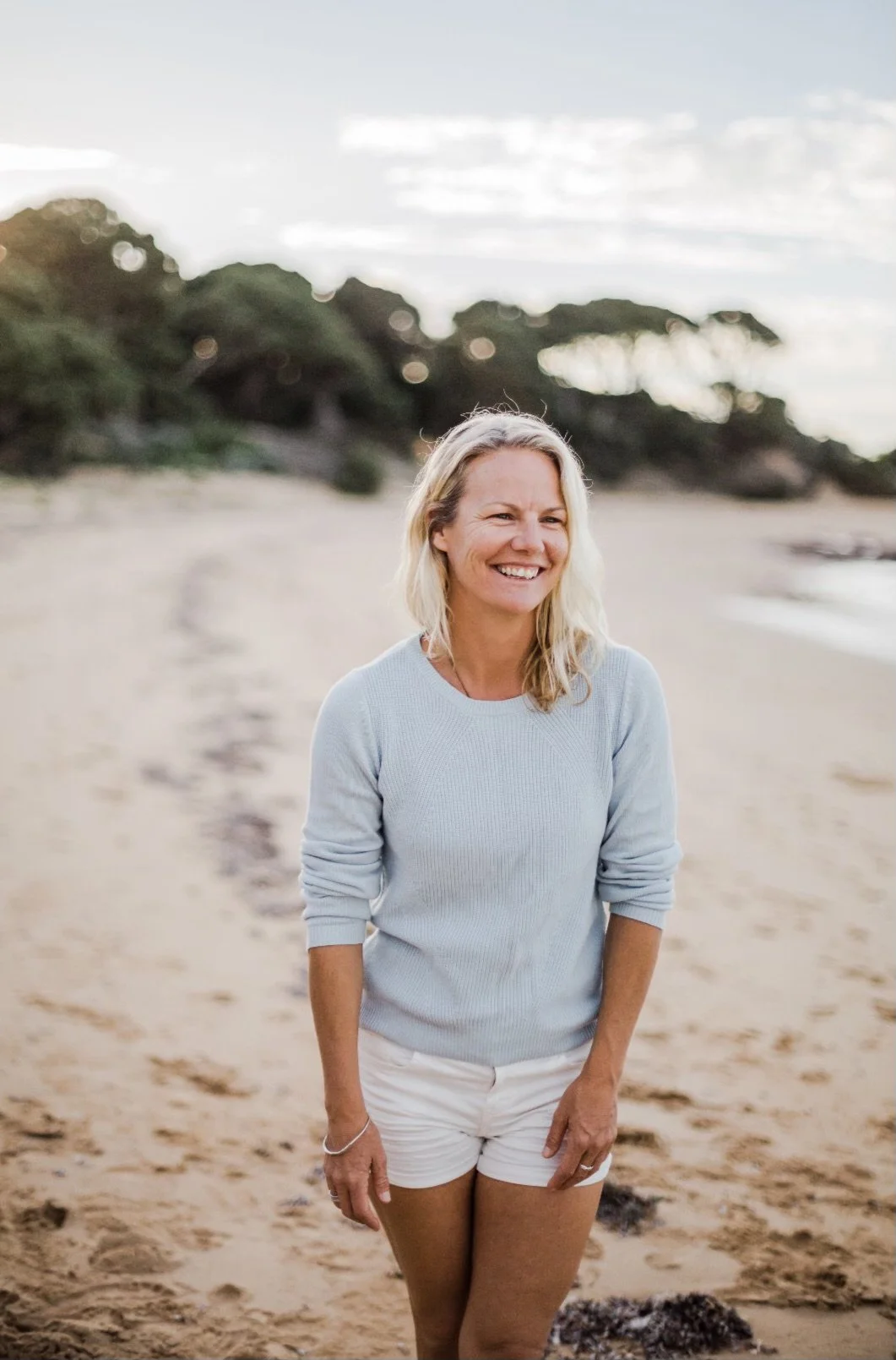 A woman with blonde hair, wearing a light blue sweater and white shorts, smiling on a sandy beach with trees and cloudy sky in the background.